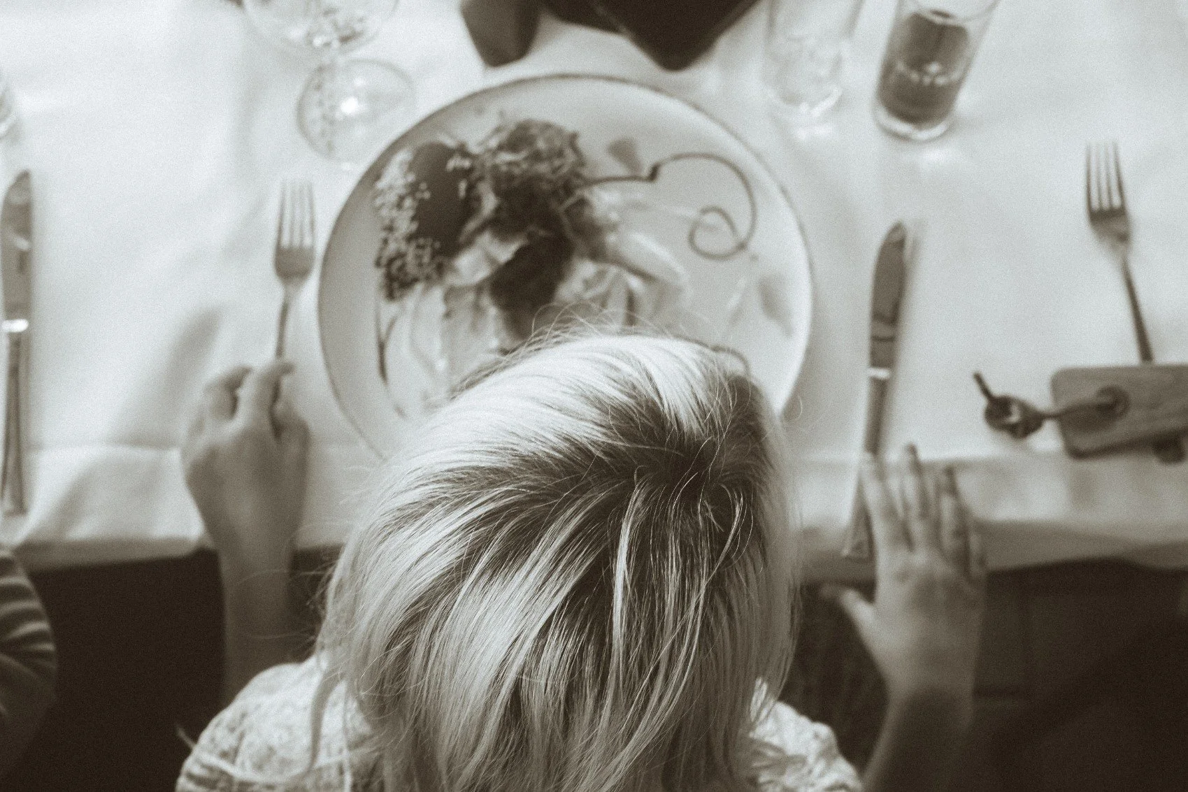 An overhead black and white shot of a person with blonde hair seated at a dining table with a plate of food, cutlery, and glasses.