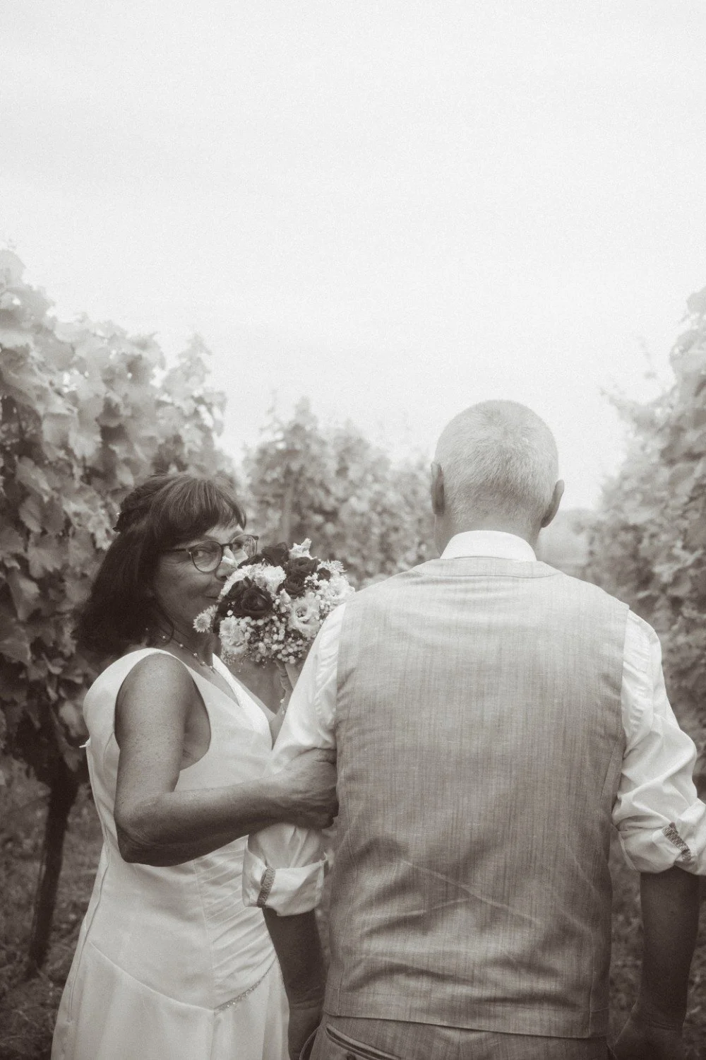 A medium black and white outdoor shot of the wedding couple walking away through a vineyard, with the bride looking back over her shoulder and smiling while holding a bouquet.