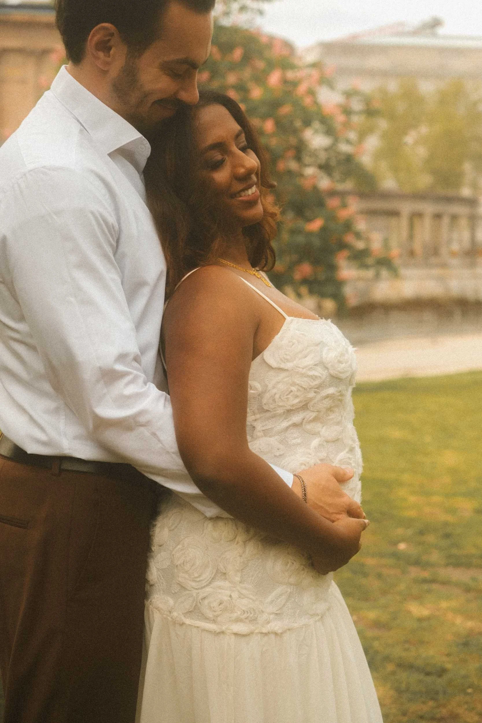 A romantic side-profile engagement portrait of a couple in Berlin, with the groom embracing the bride from behind, his hands on her stomach, as they both smile with her head tilted back against his chest.