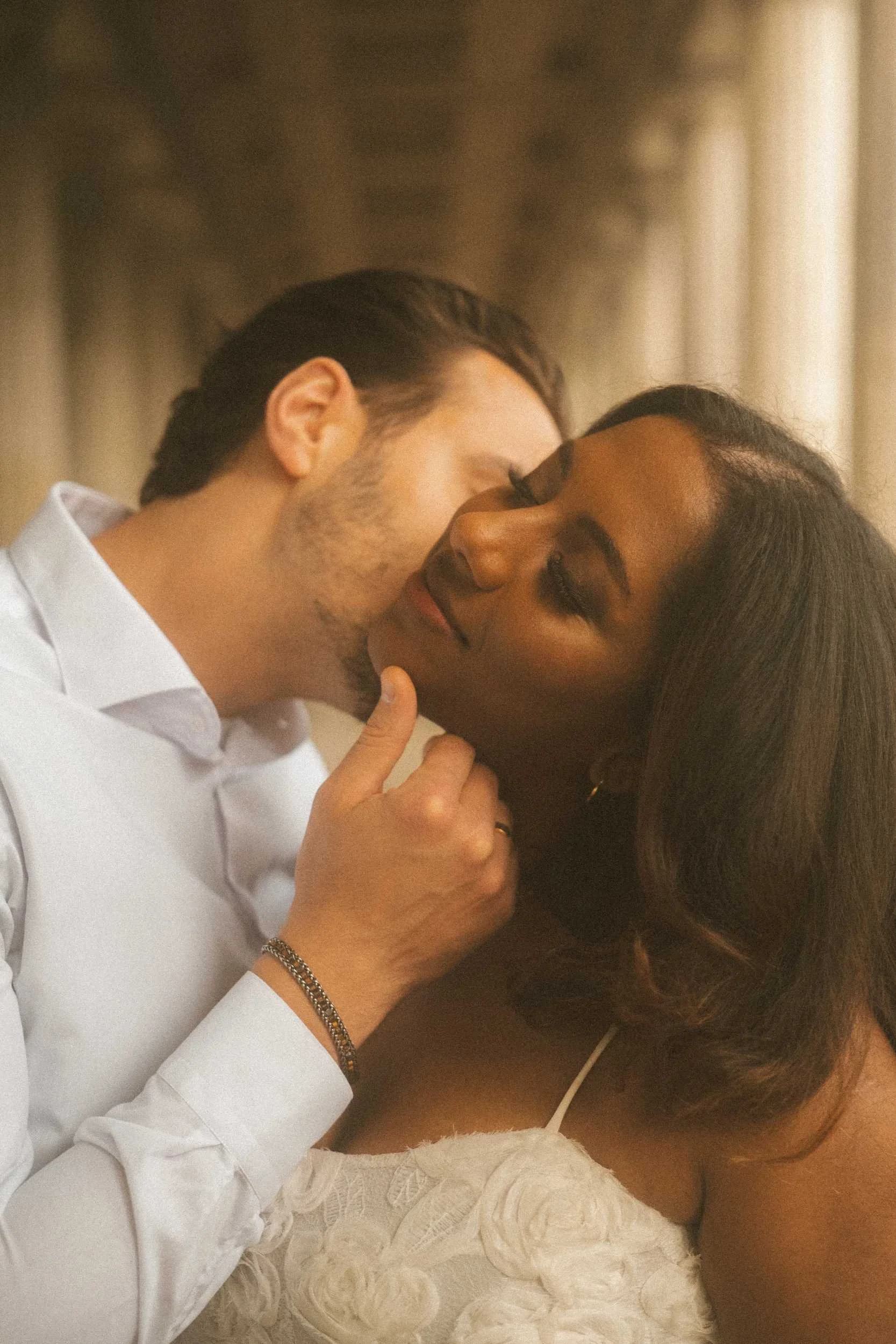 An intimate engagement portrait of a couple in Berlin, with the man gently tilting the woman's chin up to kiss her on the cheek while she smiles with her eyes closed.