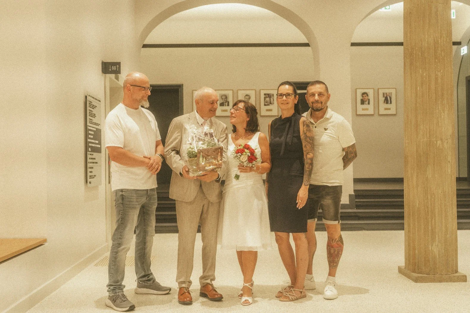 A group portrait of the bride and groom holding a bouquet and gift basket alongside guests in a bright indoor hallway.