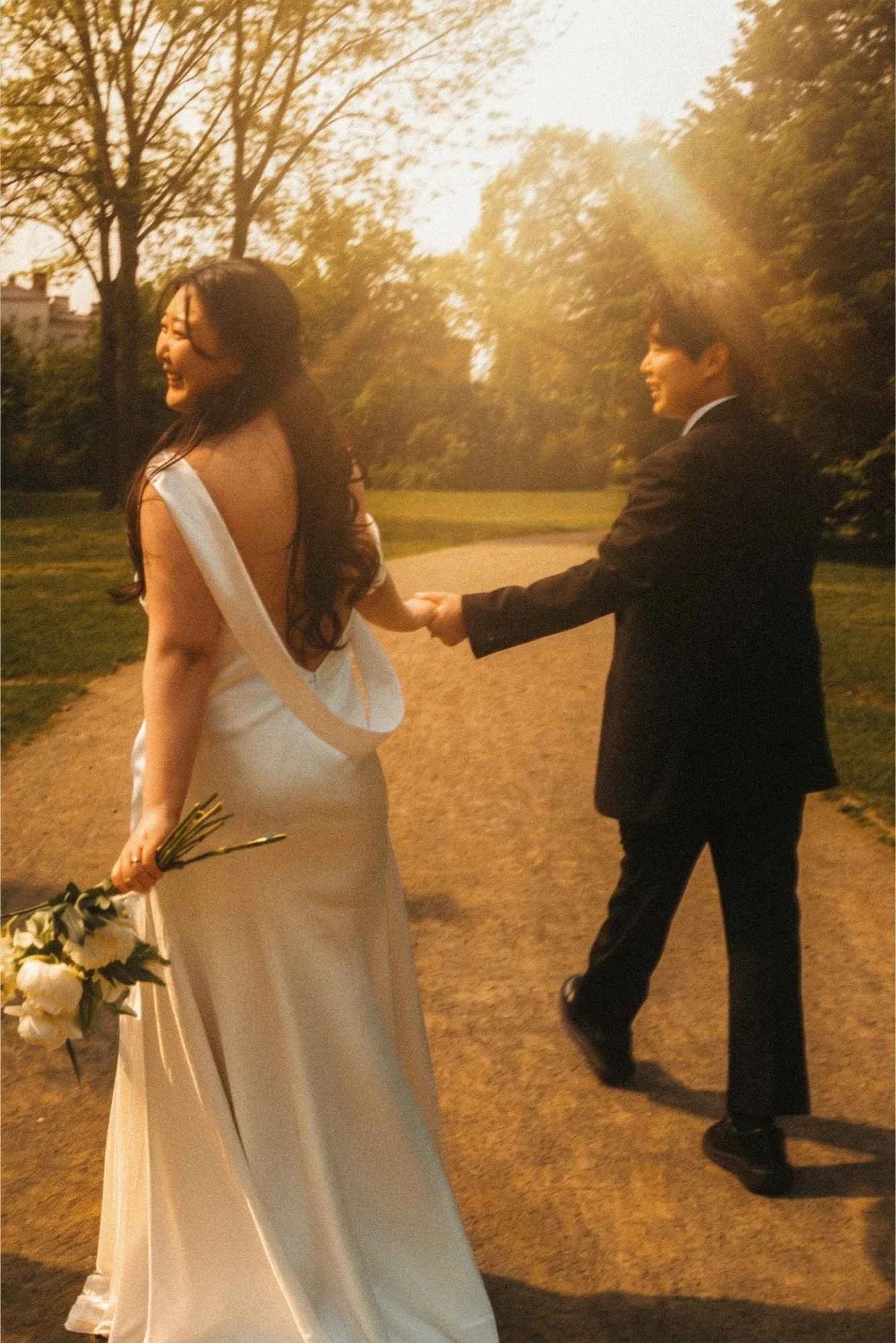 A bride and groom walking hand in hand along a path in the palace gardens in Potsdam during golden hour, with the bride looking back and smiling while the groom looks at her.