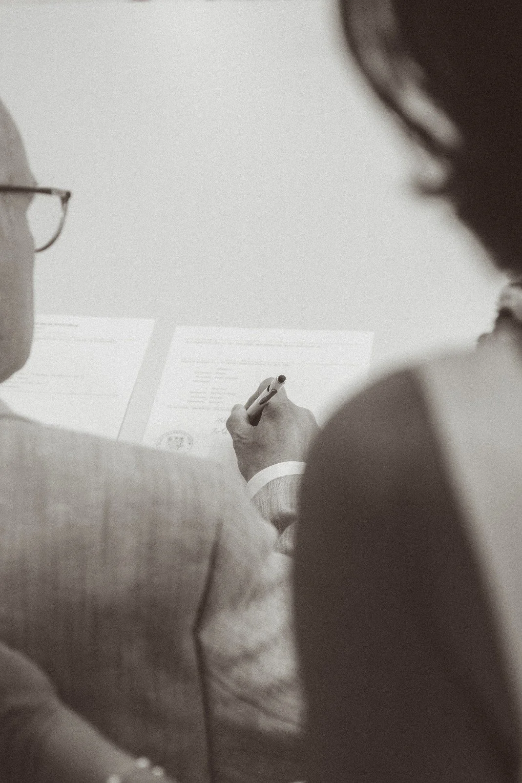 An over-the-shoulder view of a groom signing the marriage certificate document during an indoor ceremony.