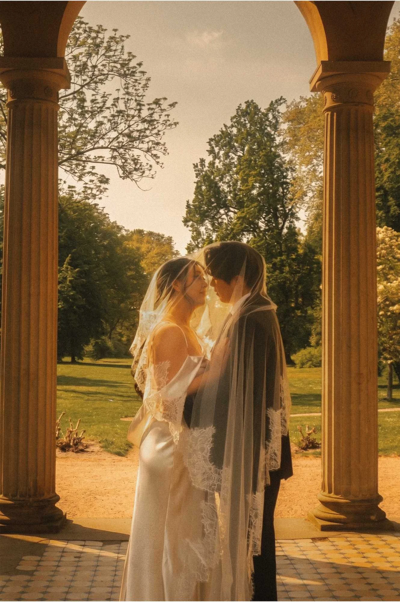 A bride and groom standing close together under a long wedding veil, framed by stone columns looking out onto a park in Potsdam.