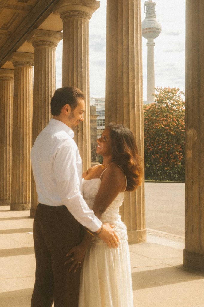 A romantic engagement portrait of a couple standing in a historic colonnade in Berlin with the iconic TV Tower in the background.