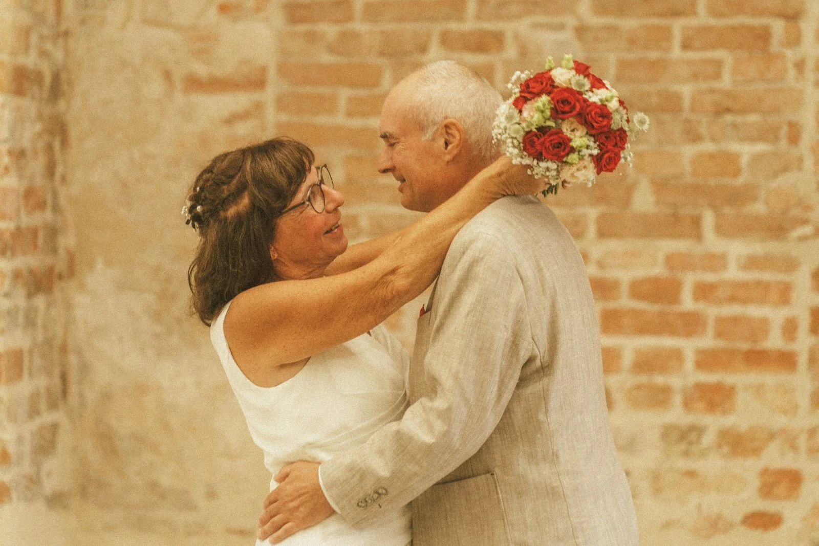 A bride holding a bouquet and embracing the groom in a warm dance in front of an exposed brick wall.
