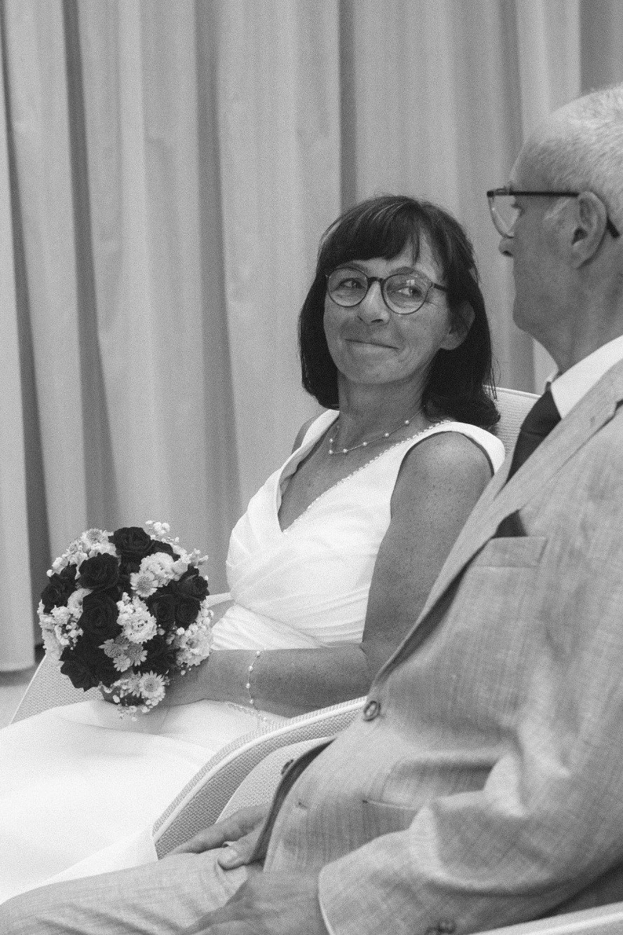 A black and white portrait of a bride holding a bouquet and smiling while seated next to the groom.