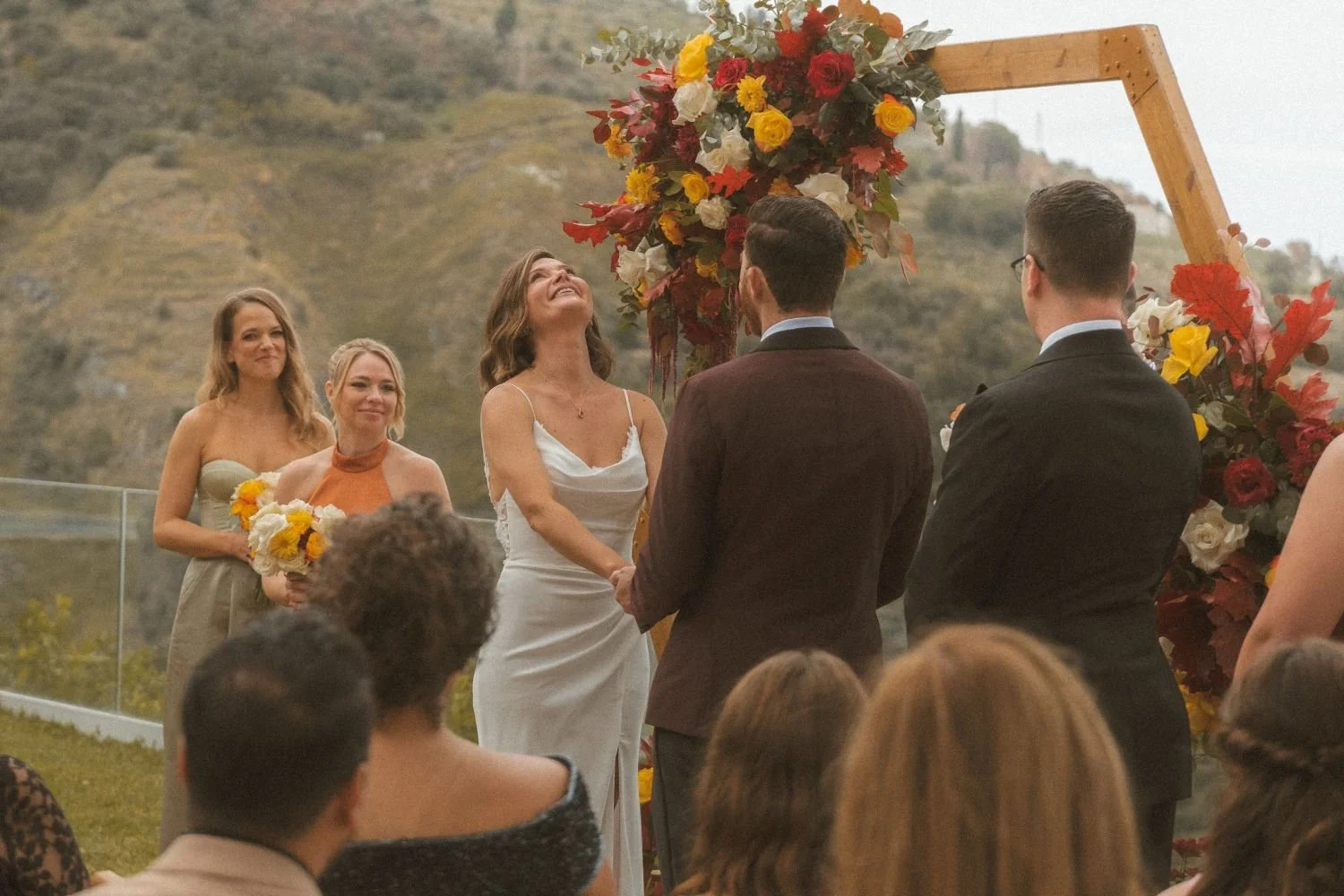 Bride and groom holding hands during an outdoor wedding ceremony beneath a floral arch with autumn colors and mountain scenery.