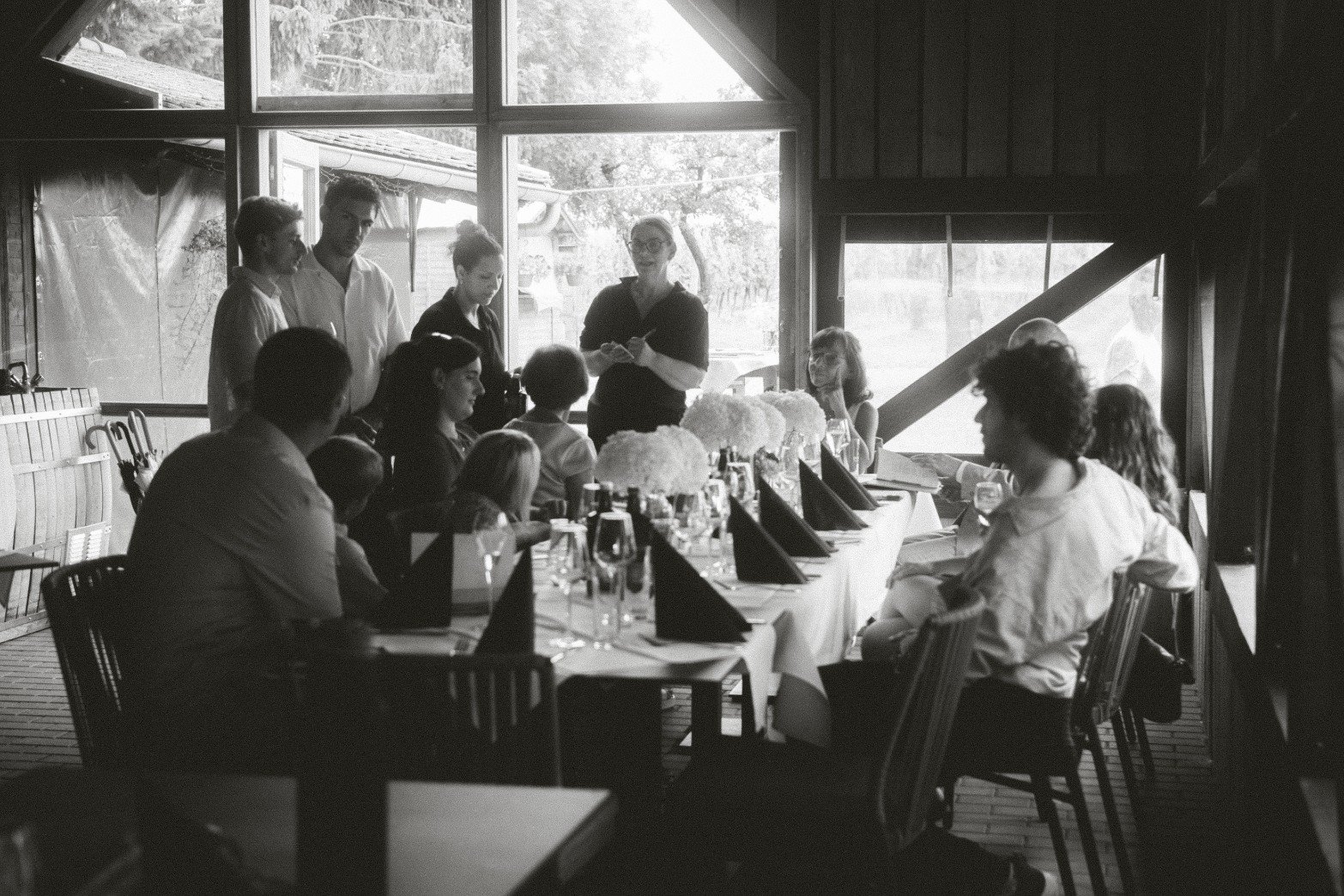 A black and white wide shot of wedding guests gathered and dining around a long table inside a venue.