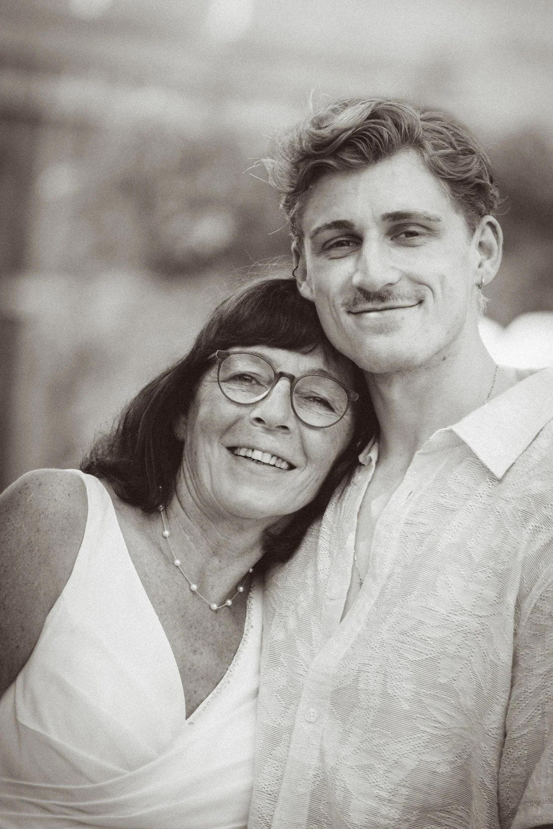 A close-up black and white portrait of a smiling woman leaning her head against a wedding guest's shoulder.