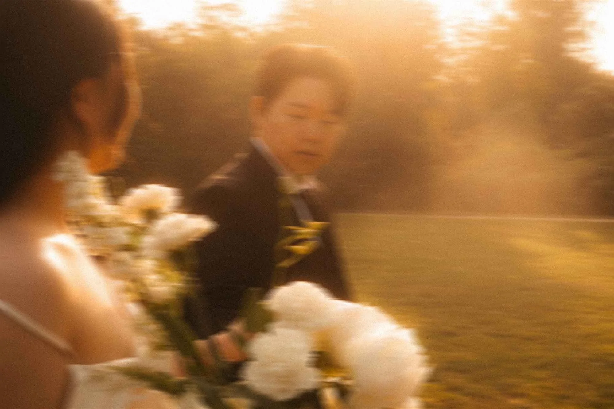 A bride in a white gown and a groom looking down and smiling while standing close together on a sunlit gravel path surrounded by trees in Potsdam.