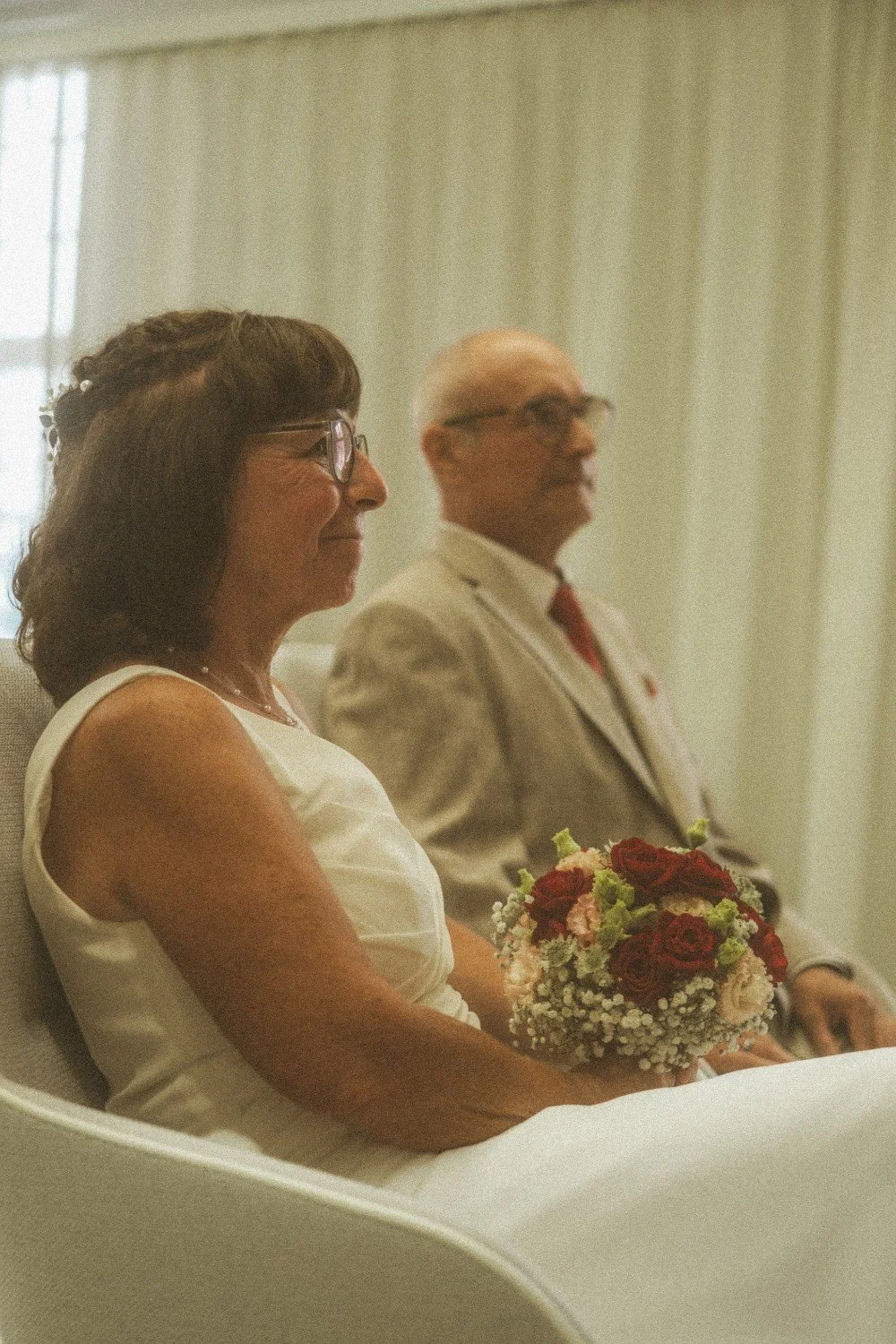 A medium indoor shot of the wedding couple seated together, with the bride holding a bouquet of red and white flowers.