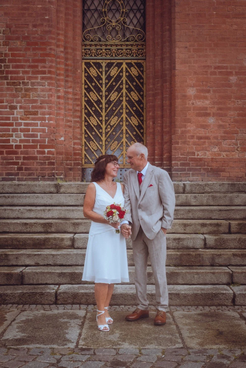 A full-length portrait of a bride holding a bouquet and holding hands with the groom while standing on stone steps in front of an ornate door and brick building.