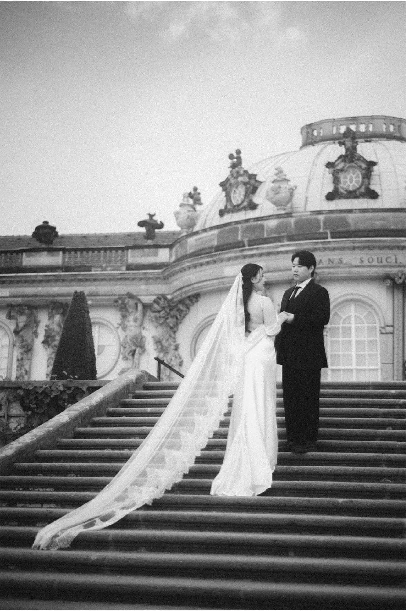 Black and white wedding portrait of a bride with a long flowing lace veil holding hands with the groom on the stone steps of Sanssouci Palace in Potsdam.