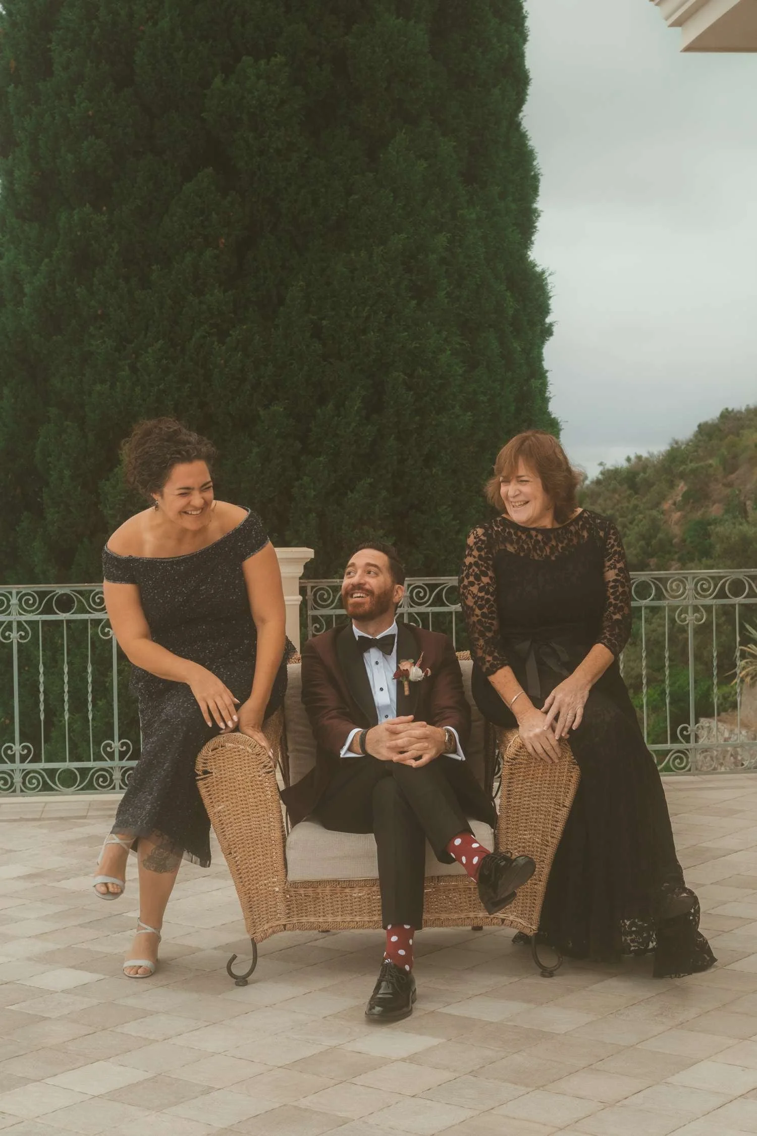 Groom sitting and laughing with two elegantly dressed women during an outdoor wedding moment on a terrace