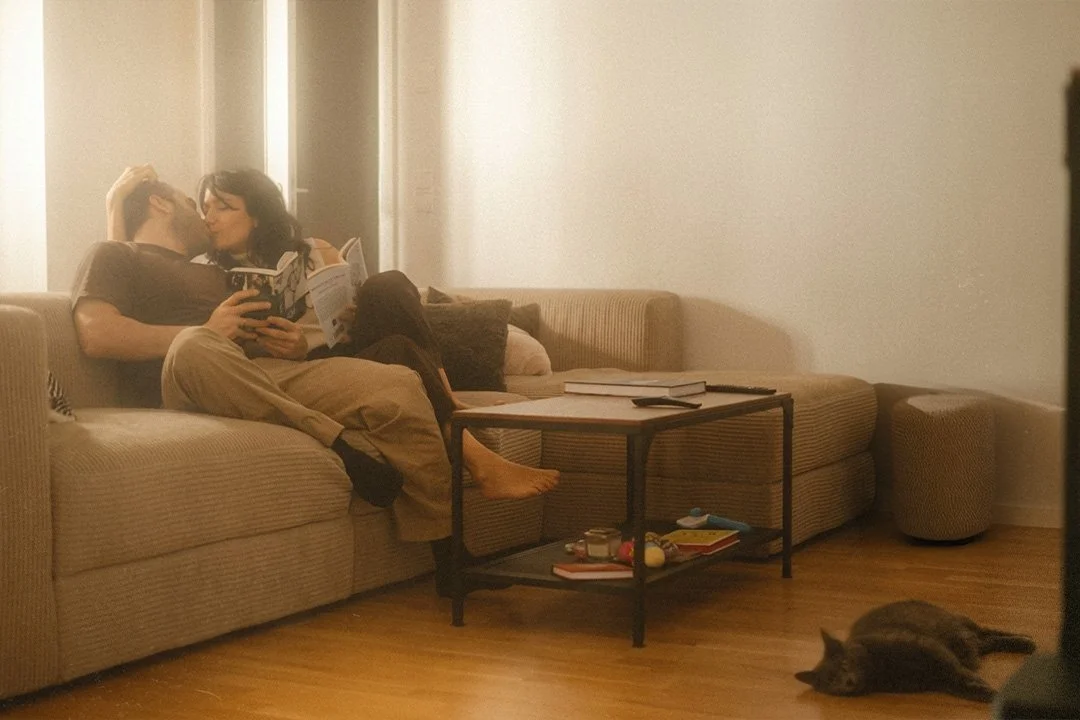Lifestyle portrait of a couple kissing on a sofa while their grey cat rests on the floor in a Berlin Kreuzberg apartment.