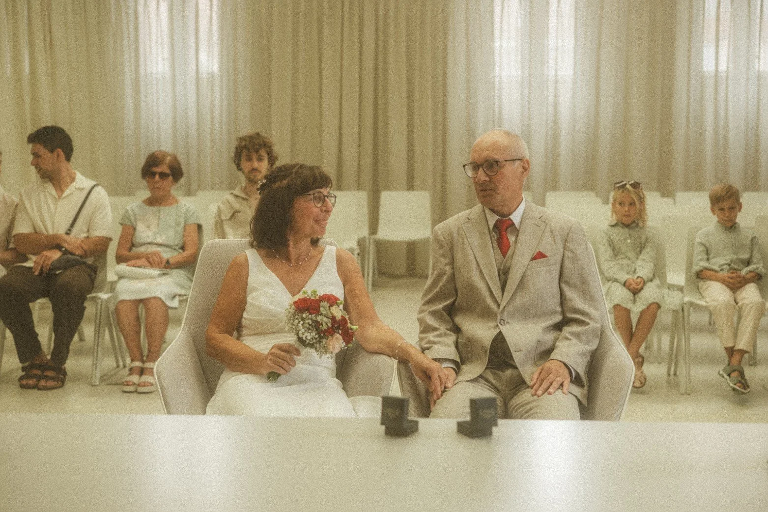 A bride and groom holding hands while seated at a table during an indoor wedding ceremony with guests watching.