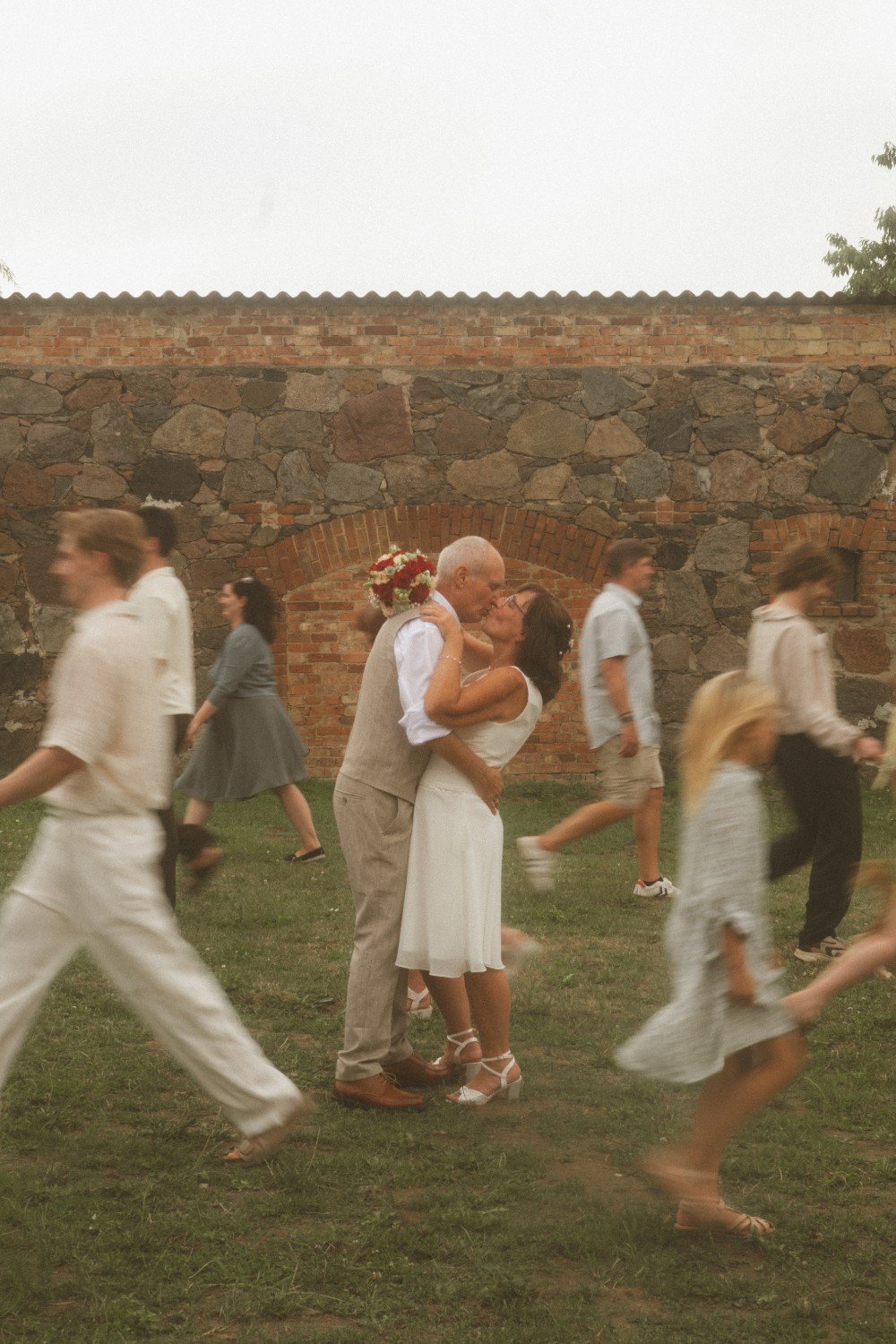 A wide outdoor shot of the wedding couple kissing in the center while guests move in blurred motion around them in front of a stone wall.