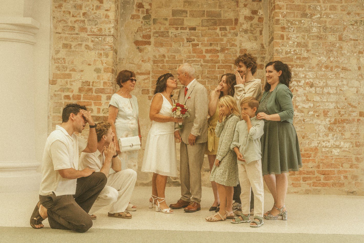 A bride and groom kissing indoors against an exposed brick wall while family members look on with playful reactions.
