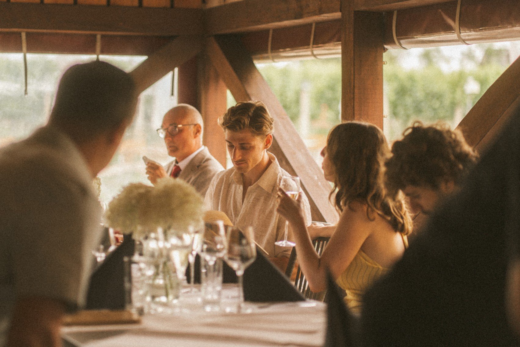 Wedding guests seated and dining together around a decorated table at an outdoor reception pavilion.