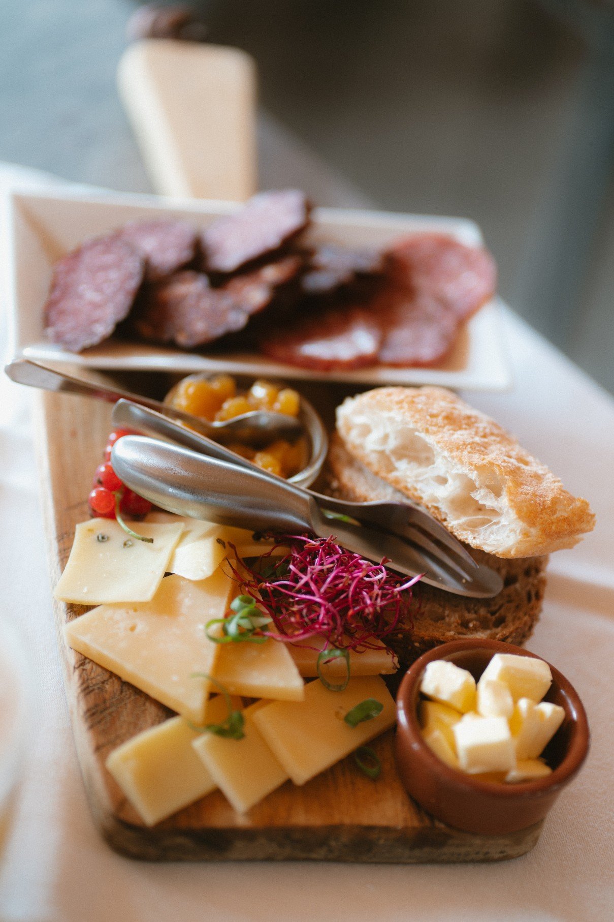 A wooden board featuring an assortment of sliced cheeses, cured meats, bread, butter, and garnishes.