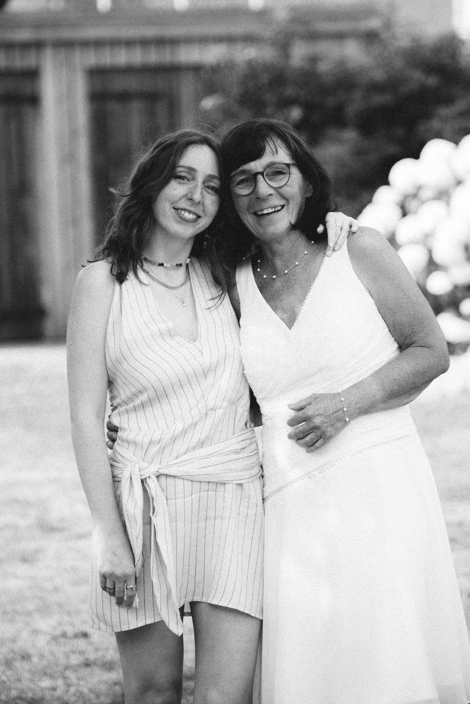 A medium black and white outdoor portrait of a smiling woman and the bride standing together with their arms around each other.