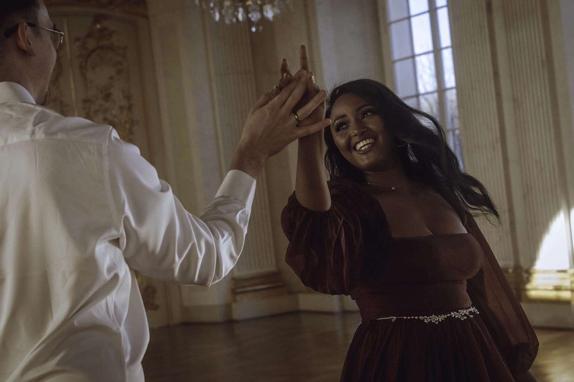 A candid moment capturing an engaged couple sharing a joyous dance inside the ornate halls of Charlottenburg Palace in Berlin. The bride-to-be radiates happiness in a flowing maroon gown with delicate embellishments, while her partner complements the