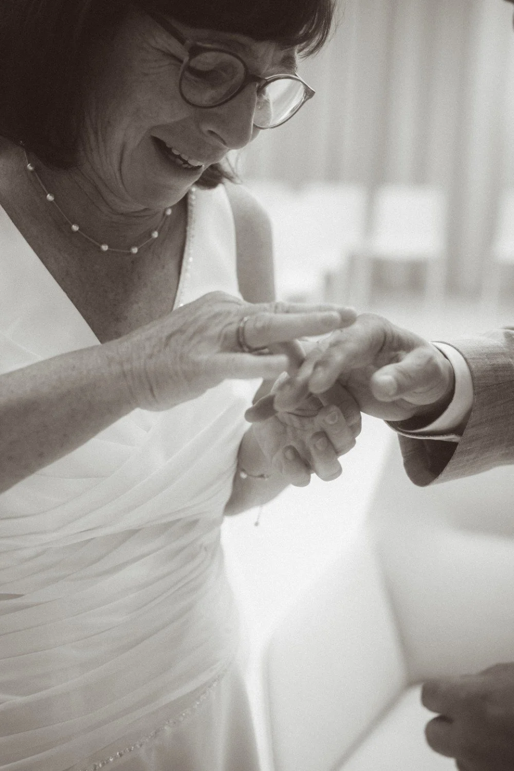 A close-up black and white indoor shot of the smiling bride placing a wedding ring onto the groom's finger.
