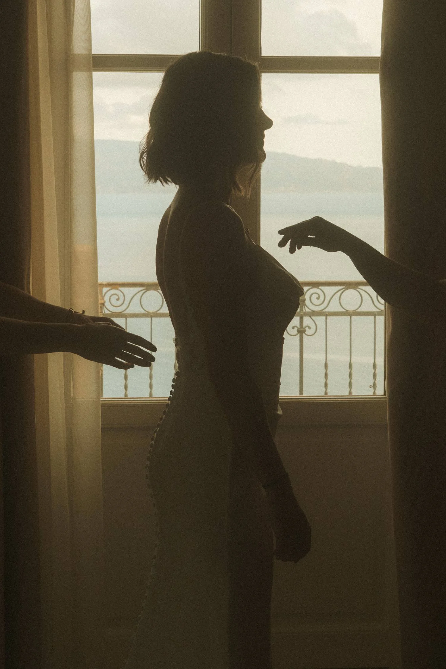 Silhouette of bride standing by window as helpers adjust wedding dress with sea view in background