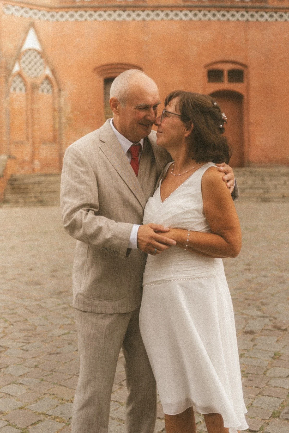 A newlywed couple embracing affectionately in a historic cobblestone square with traditional buildings in the background.