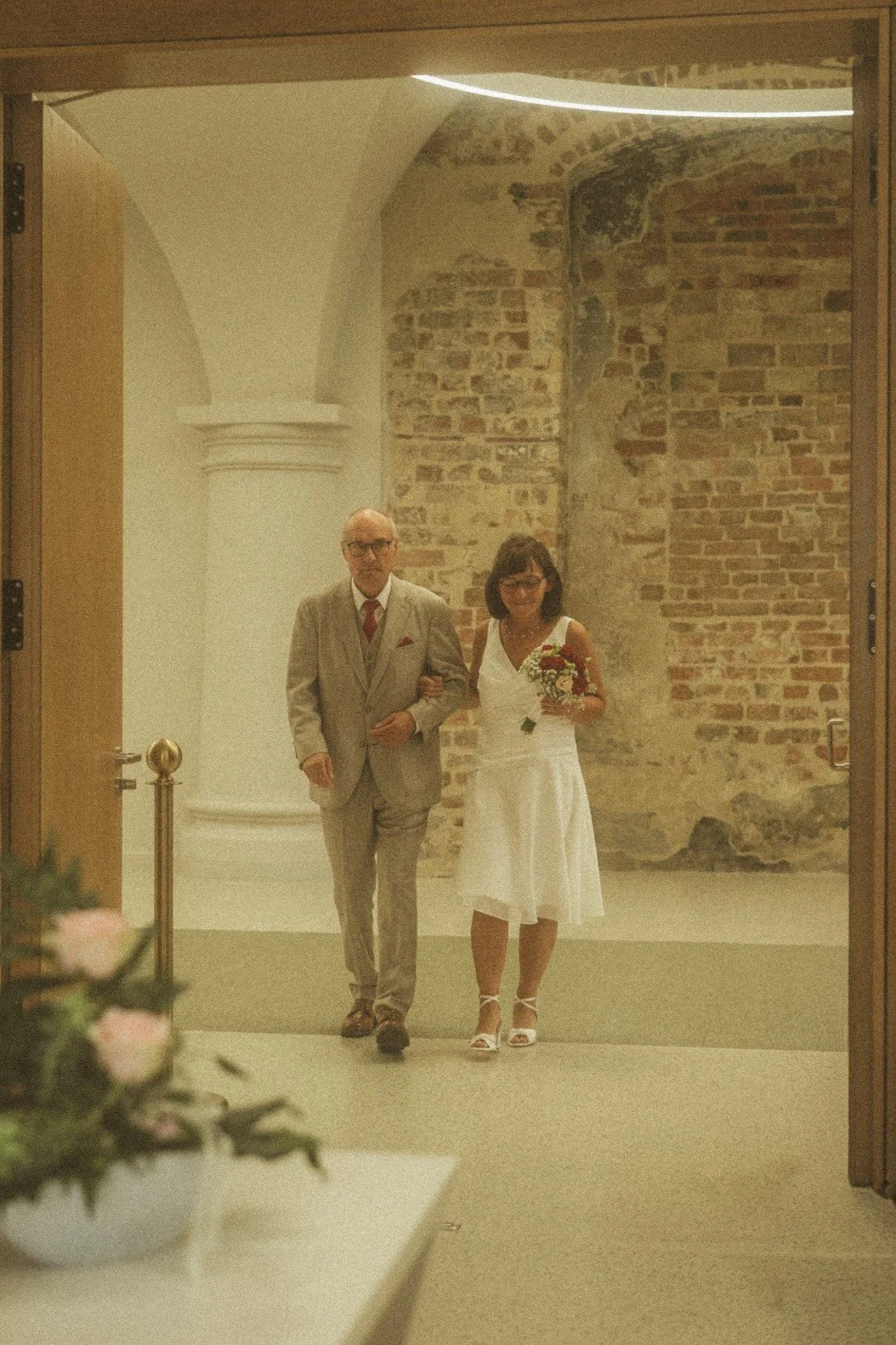 A bride holding a bouquet and walking arm-in-arm with the groom through an indoor doorway.