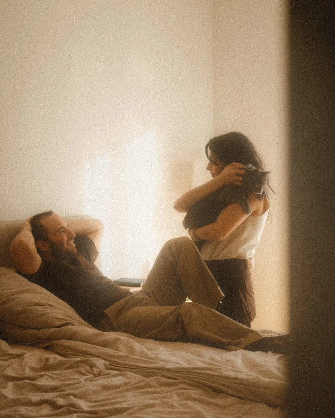 Lifestyle portrait of a man lying on a bed while a woman stands holding and petting a grey cat in a Berlin Kreuzberg apartment.