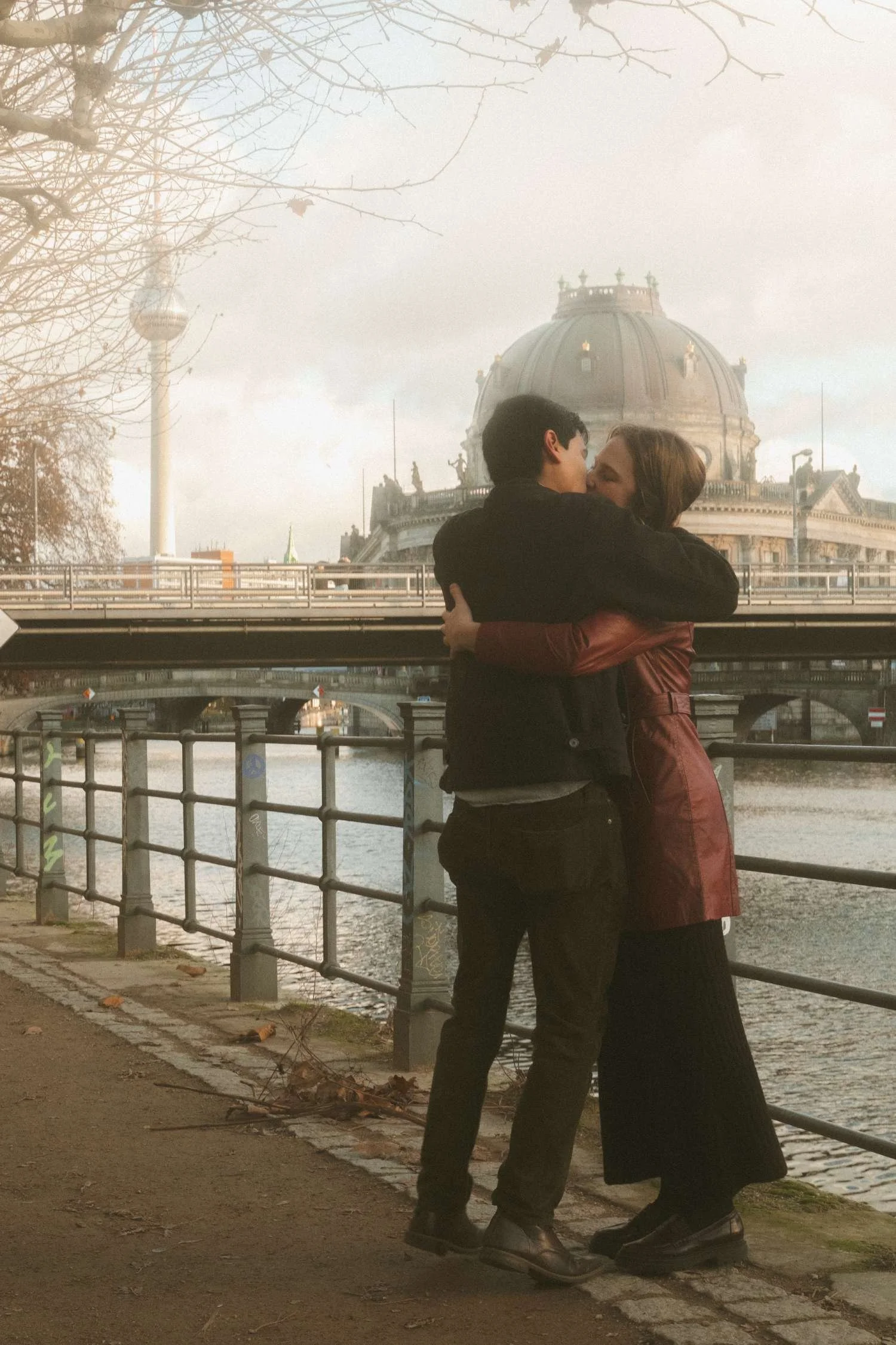 Couple kissing by the Spree River with Berliner Dom and Fernsehturm in Berlin during a cinematic winter photoshoot.