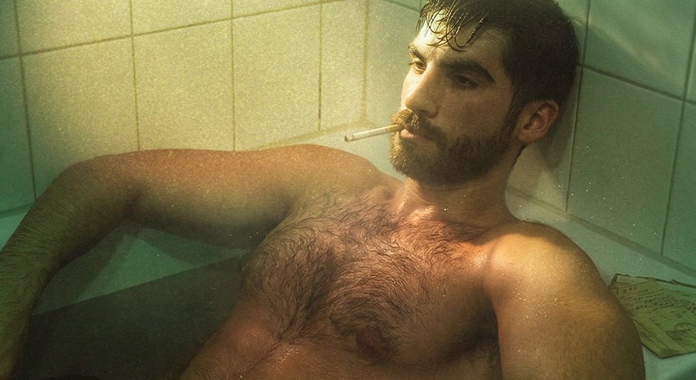 A bearded man relaxing in a tiled bathtub with a cigarette in his mouth under warm, cinematic lighting in Berlin.