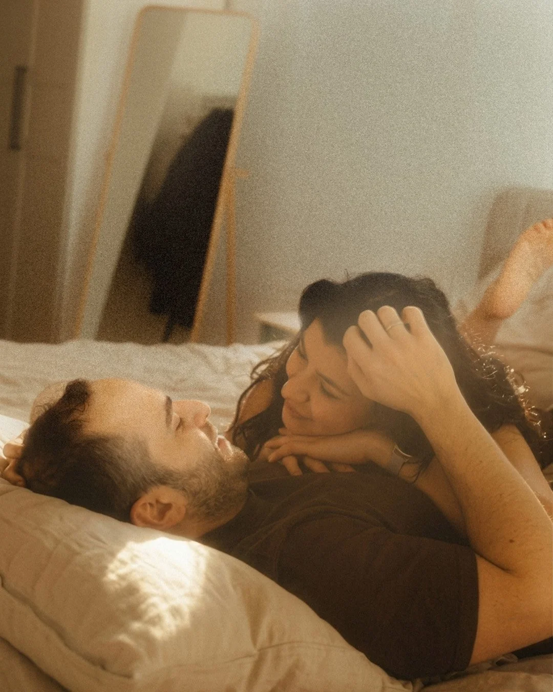 Intimate lifestyle portrait of a couple lying in bed and looking at each other in a warm, sunlit Berlin Kreuzberg apartment.