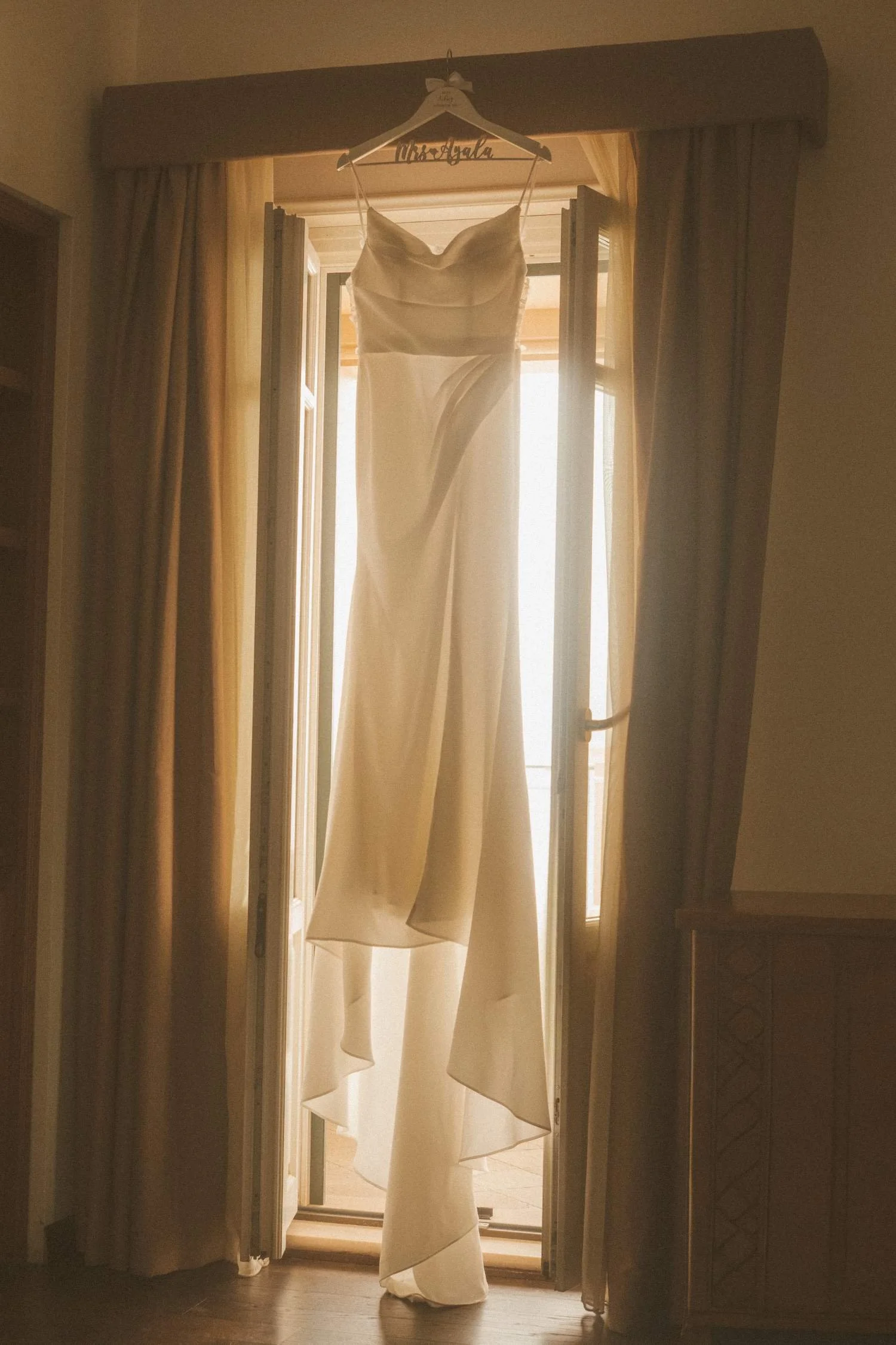 Elegant wedding dress hanging in front of an open window, softly backlit during bridal wedding preparations.