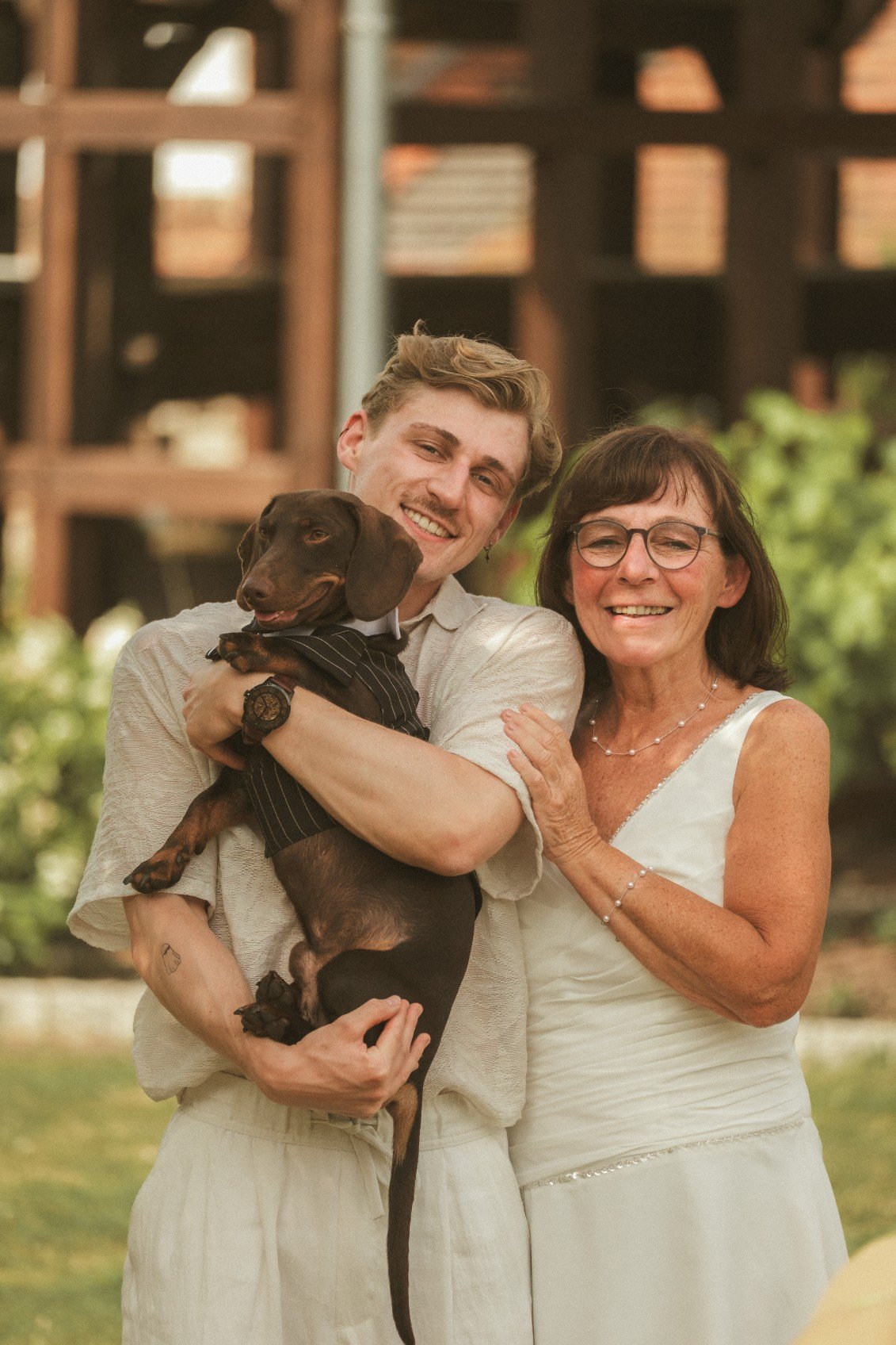 A close-up outdoor portrait of a young man holding a small dog in a tiny suit, standing next to the smiling bride in a white dress.