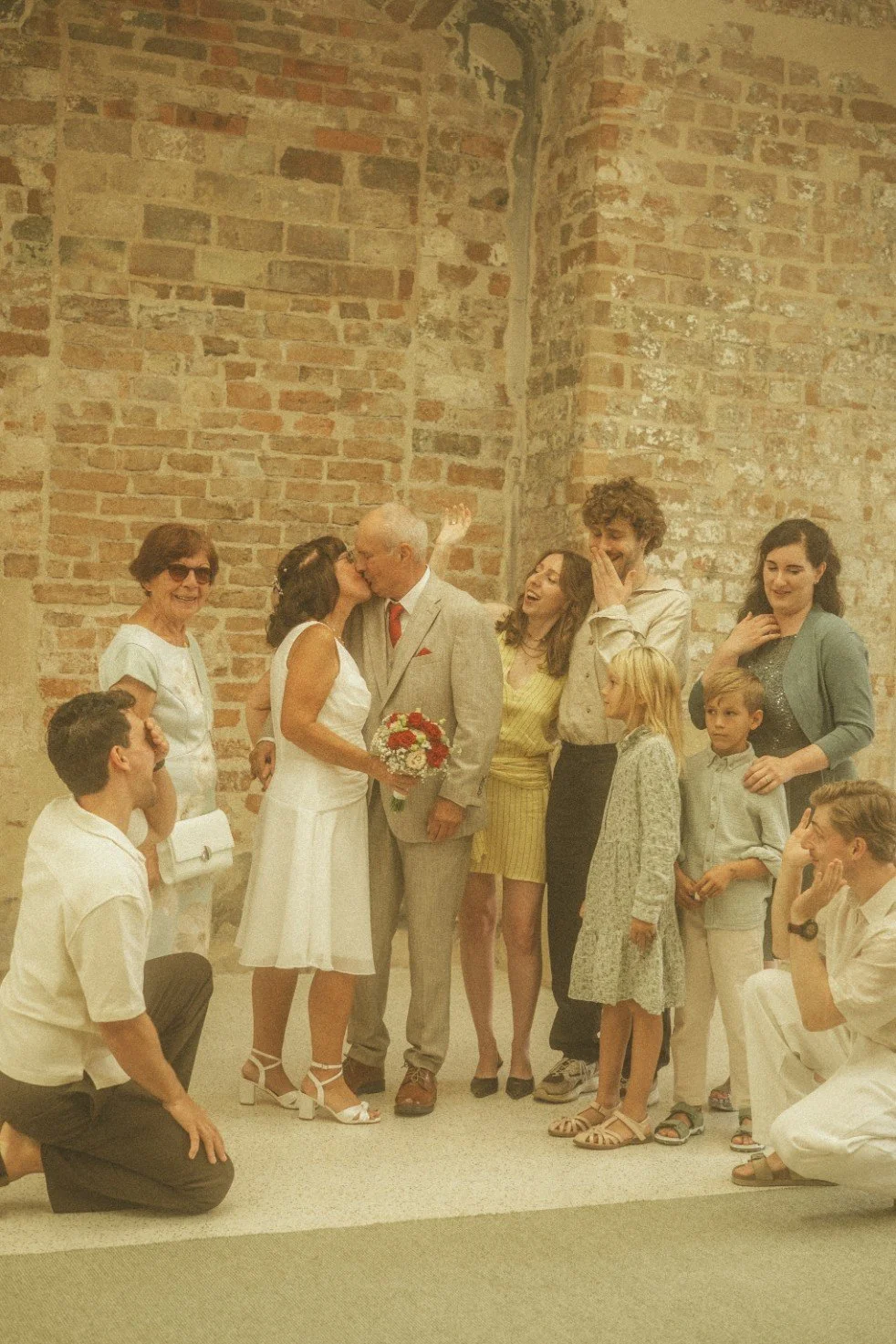 A group portrait of the wedding couple kissing while guests react and smile around them in front of a rustic brick wall.