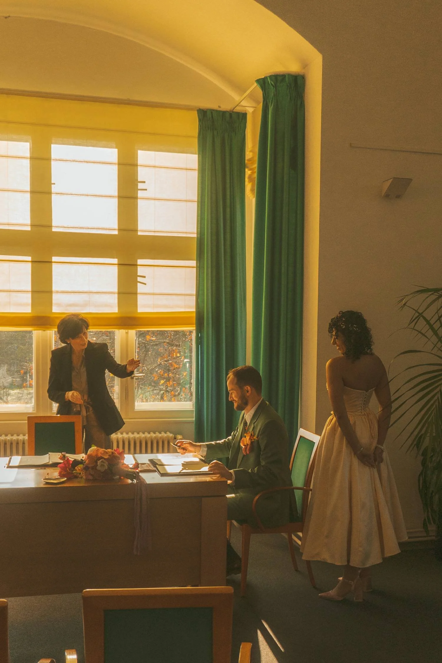 Groom signing official documents during a civil wedding ceremony at the Berlin civil registry office while the bride waits nearby.