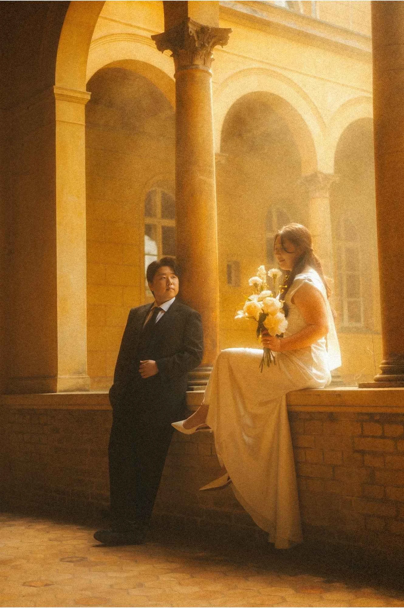 A bride holding a white floral bouquet sits on a stone ledge while the groom stands leaning beside her, framed by the sunlit arches and columns of an ancient cloister courtyard in Potsdam.