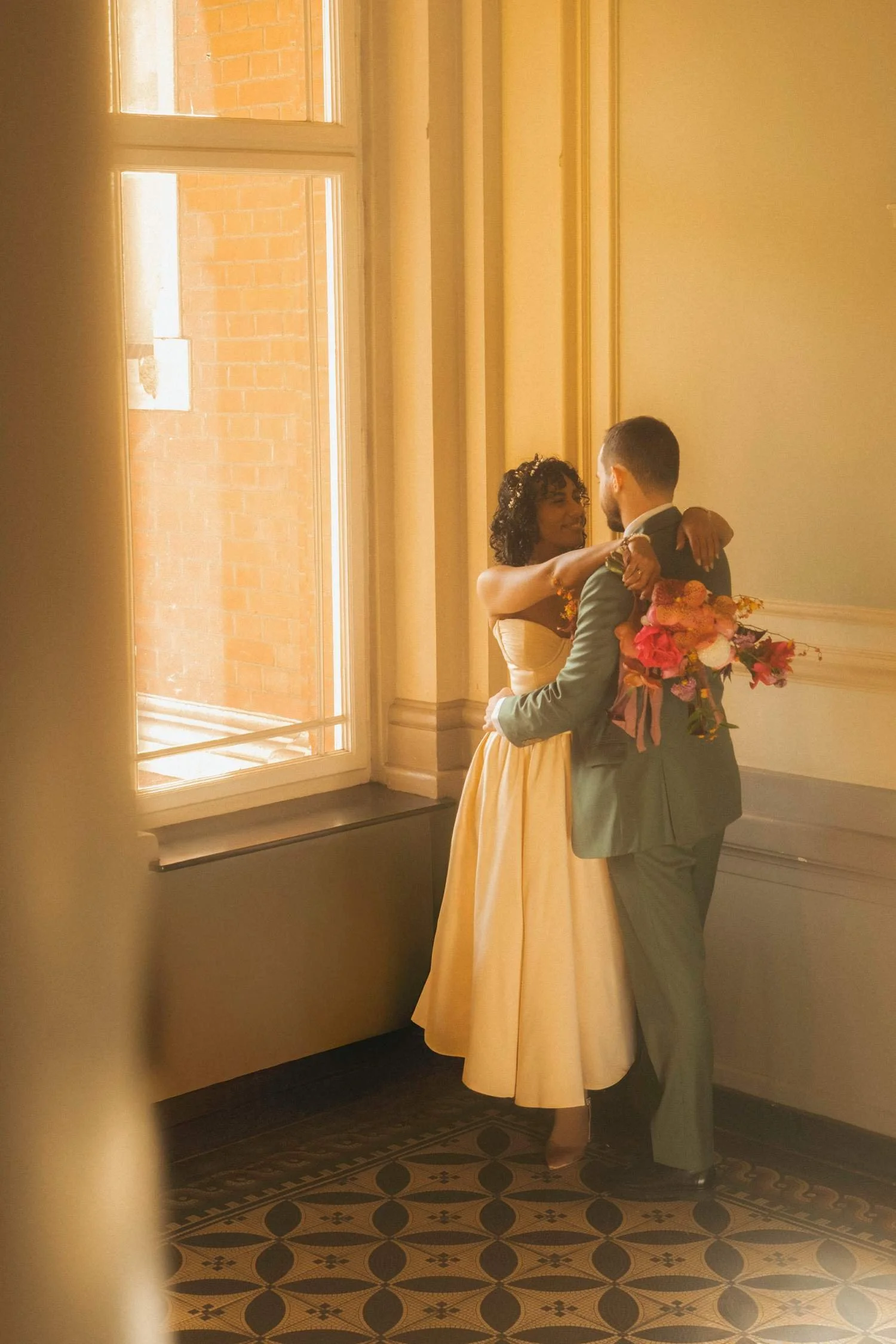 Bride and groom holding each other in soft window light, sharing a quiet moment indoors after the ceremony.