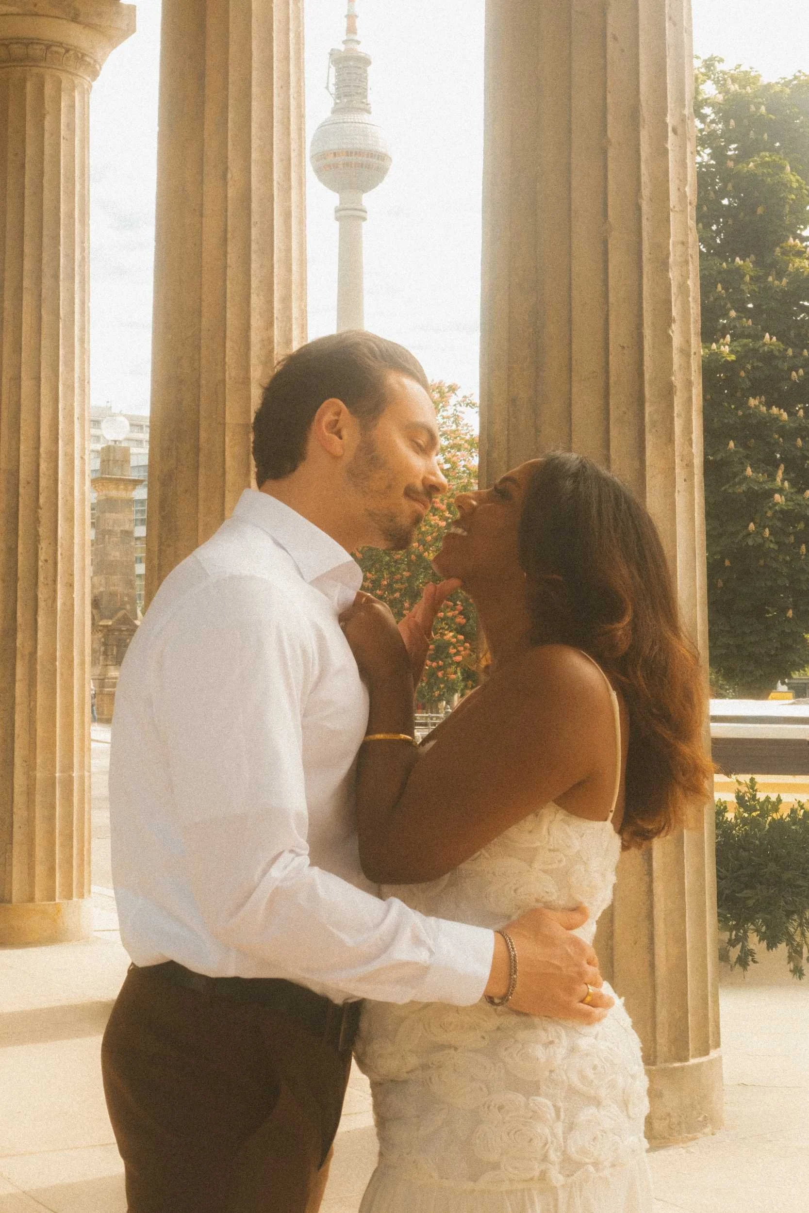 A romantic engagement portrait of a couple standing between historic neoclassical columns in Berlin, staring into each other's eyes and smiling with the TV Tower in the background.