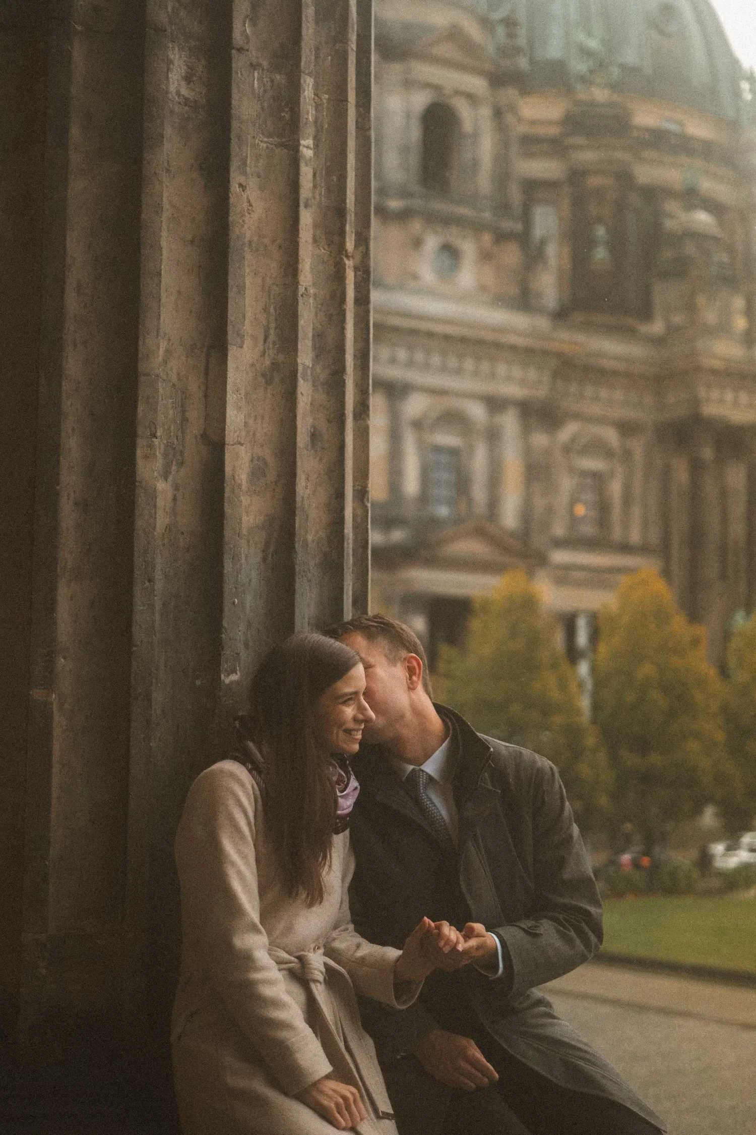 Couple sharing intimate moment by stone columns with Berliner Dom in background during Berlin photoshoot