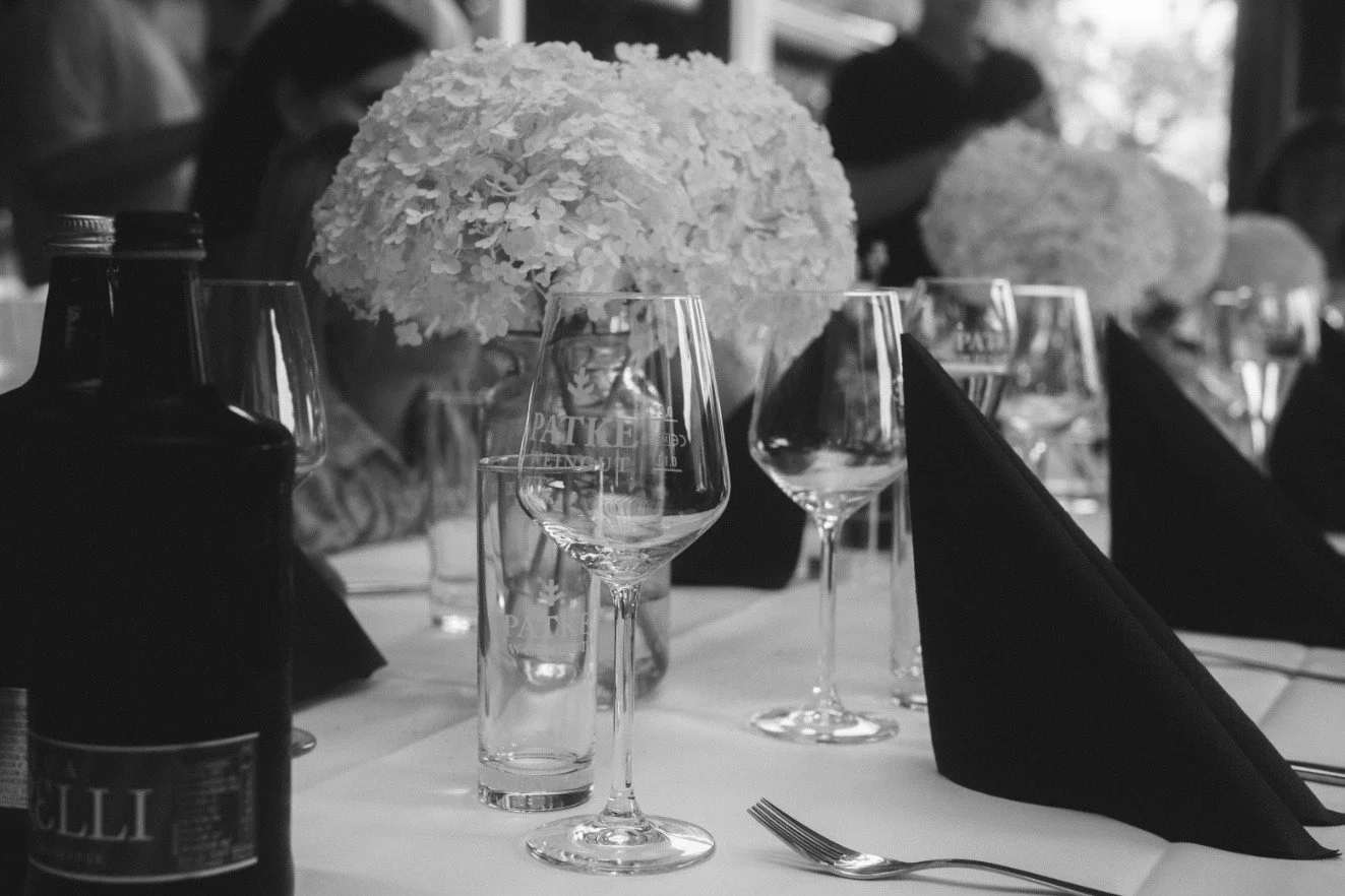 A black and white close-up of a wedding table setting featuring wine glasses, napkins, bottles, and floral centerpieces.