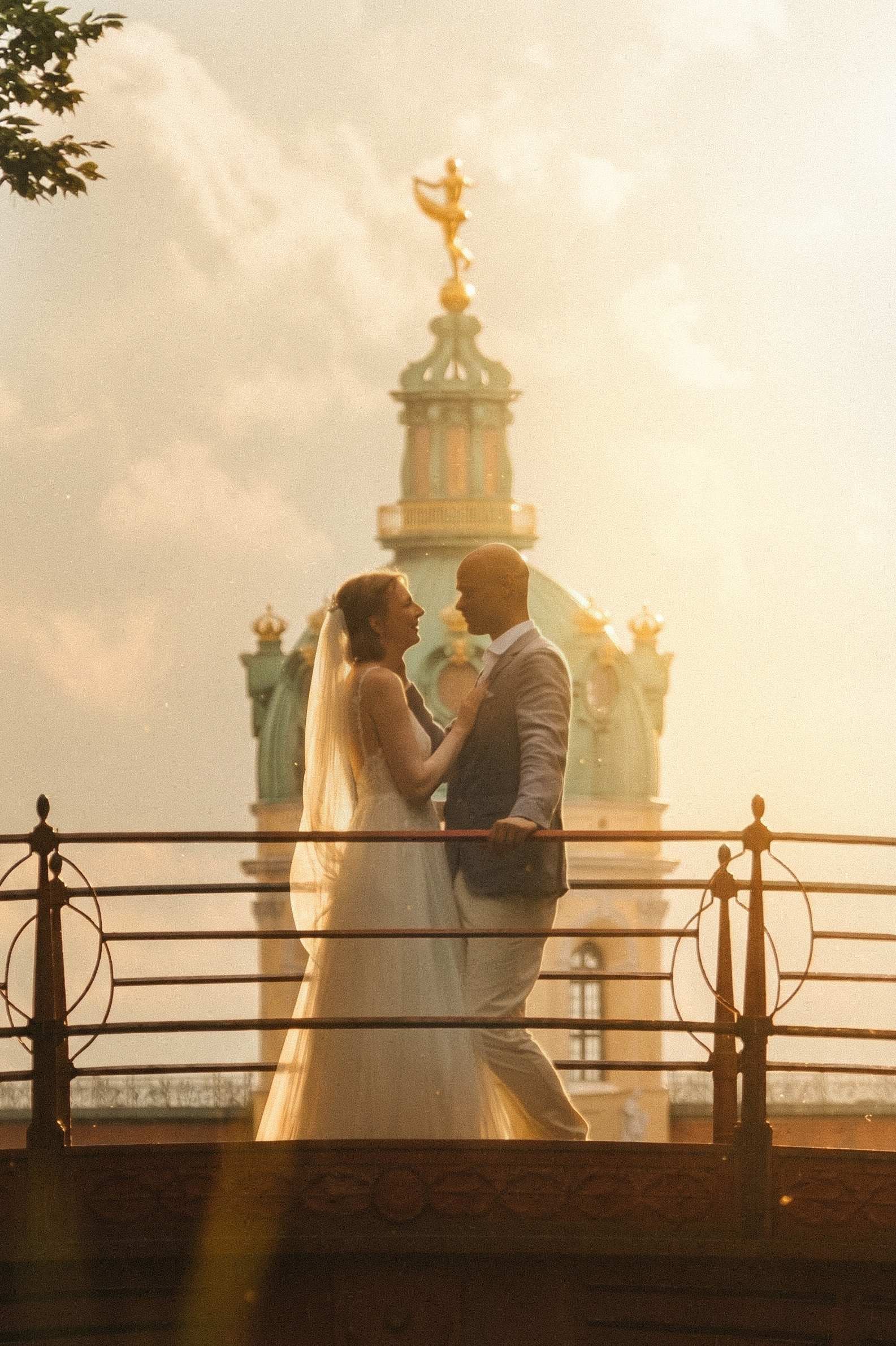 Newlyweds on ornate bridge at golden hour with Charlottenburg Belvedere dome and golden statue, Berlin