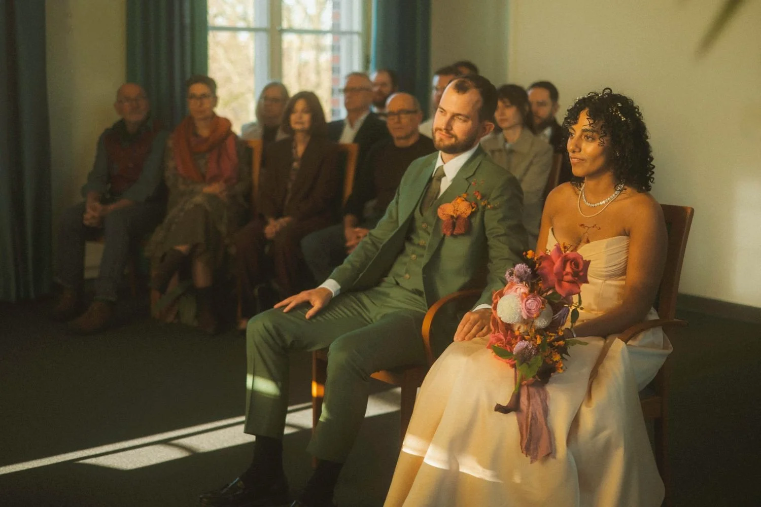 Bride and groom seated together during a wedding ceremony at a registry office, with guests seated behind them.