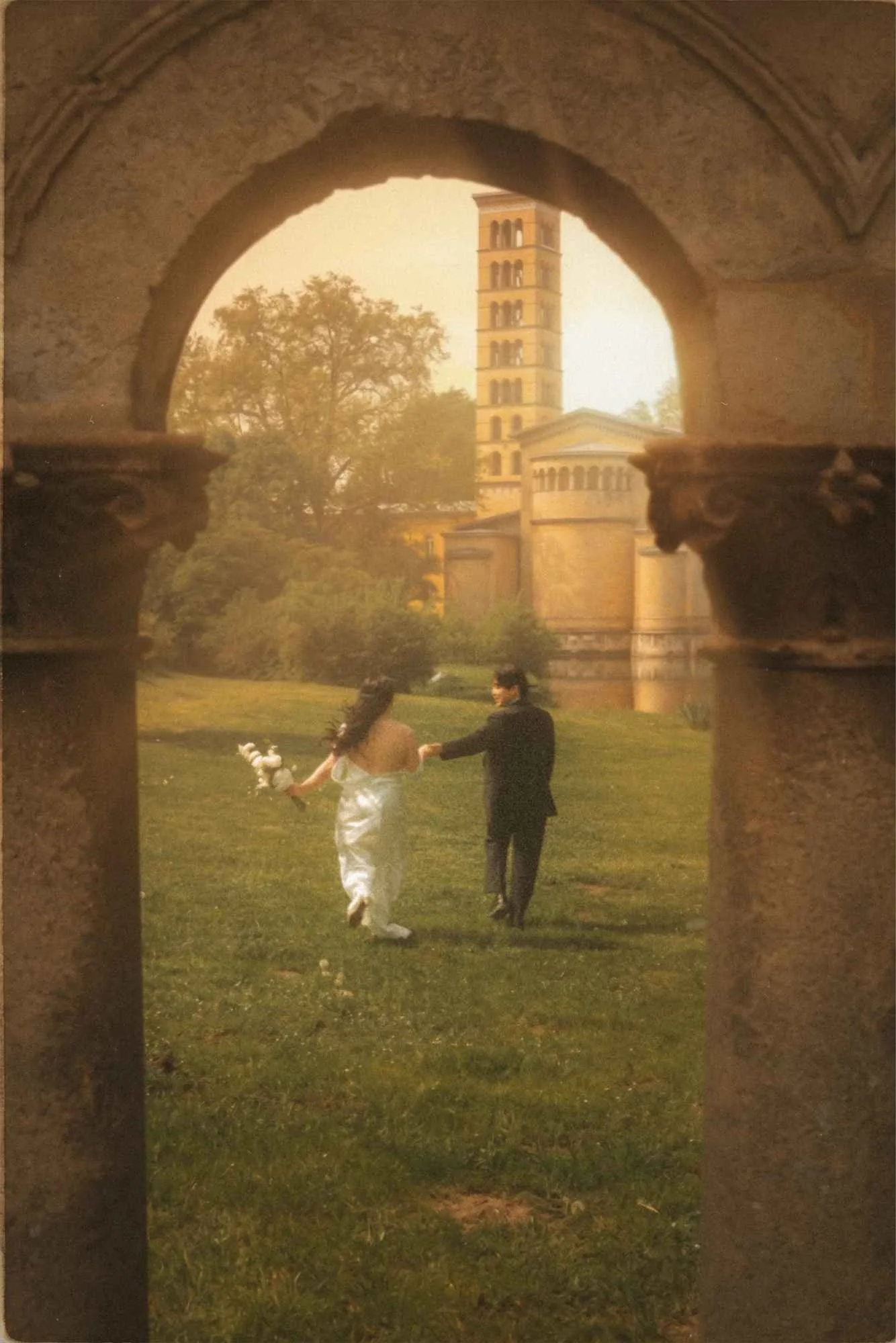 A wide-angle shot of a bride and groom holding hands and running through a field, perfectly framed between the large architectural stone columns of a palace in Potsdam during golden hour.