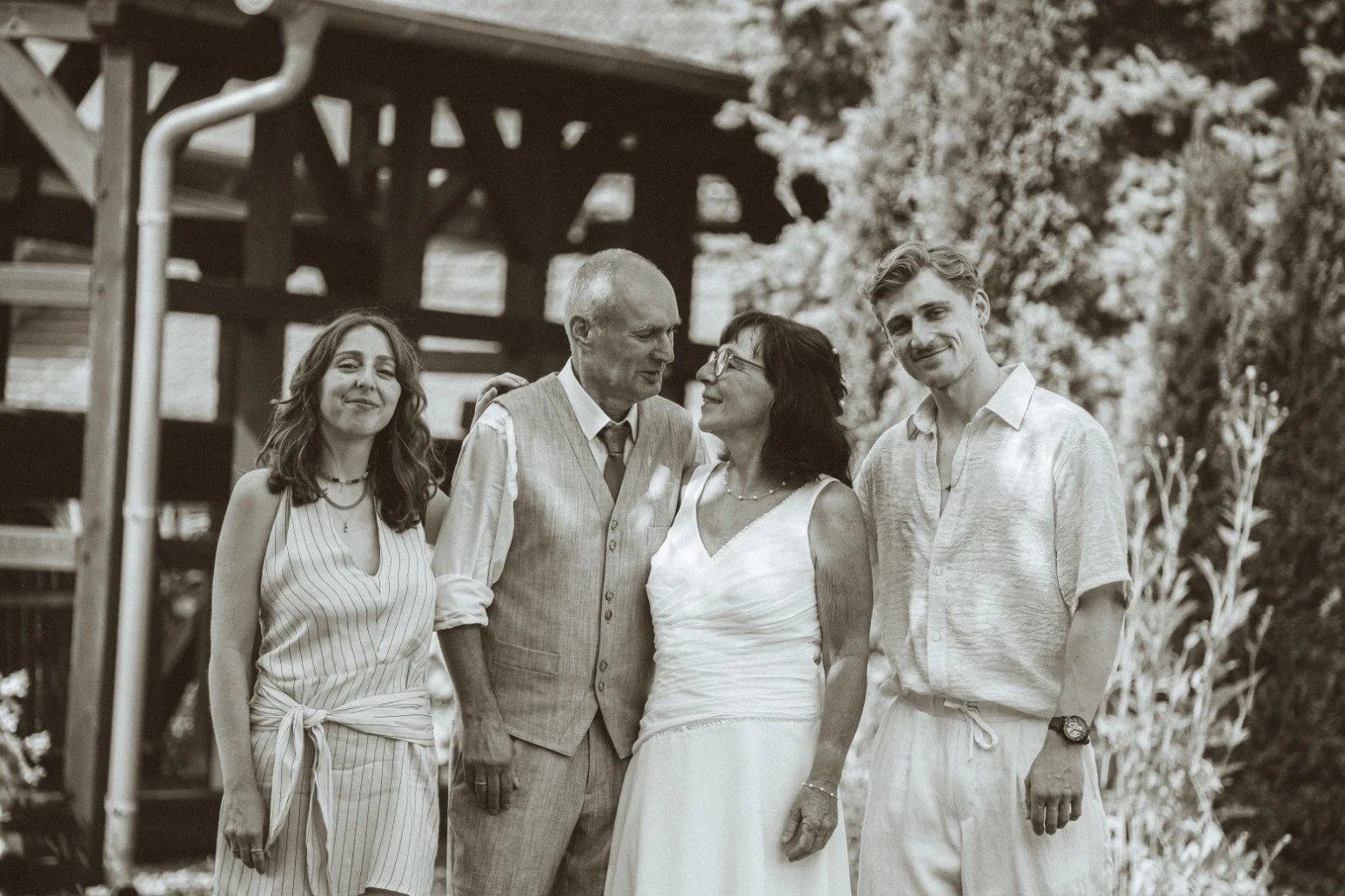 A medium black and white group portrait of the wedding couple posing outdoors alongside a man and a woman near a wooden structure.