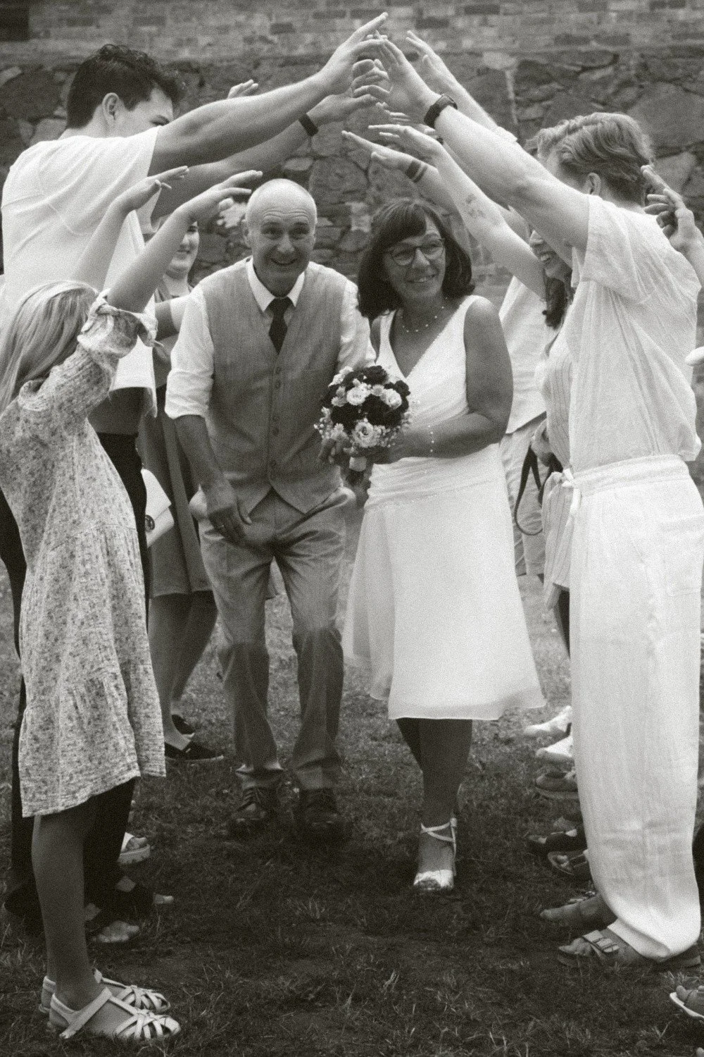 A medium black and white outdoor shot of the smiling wedding couple walking underneath an arch formed by the raised arms and hands of their guests.