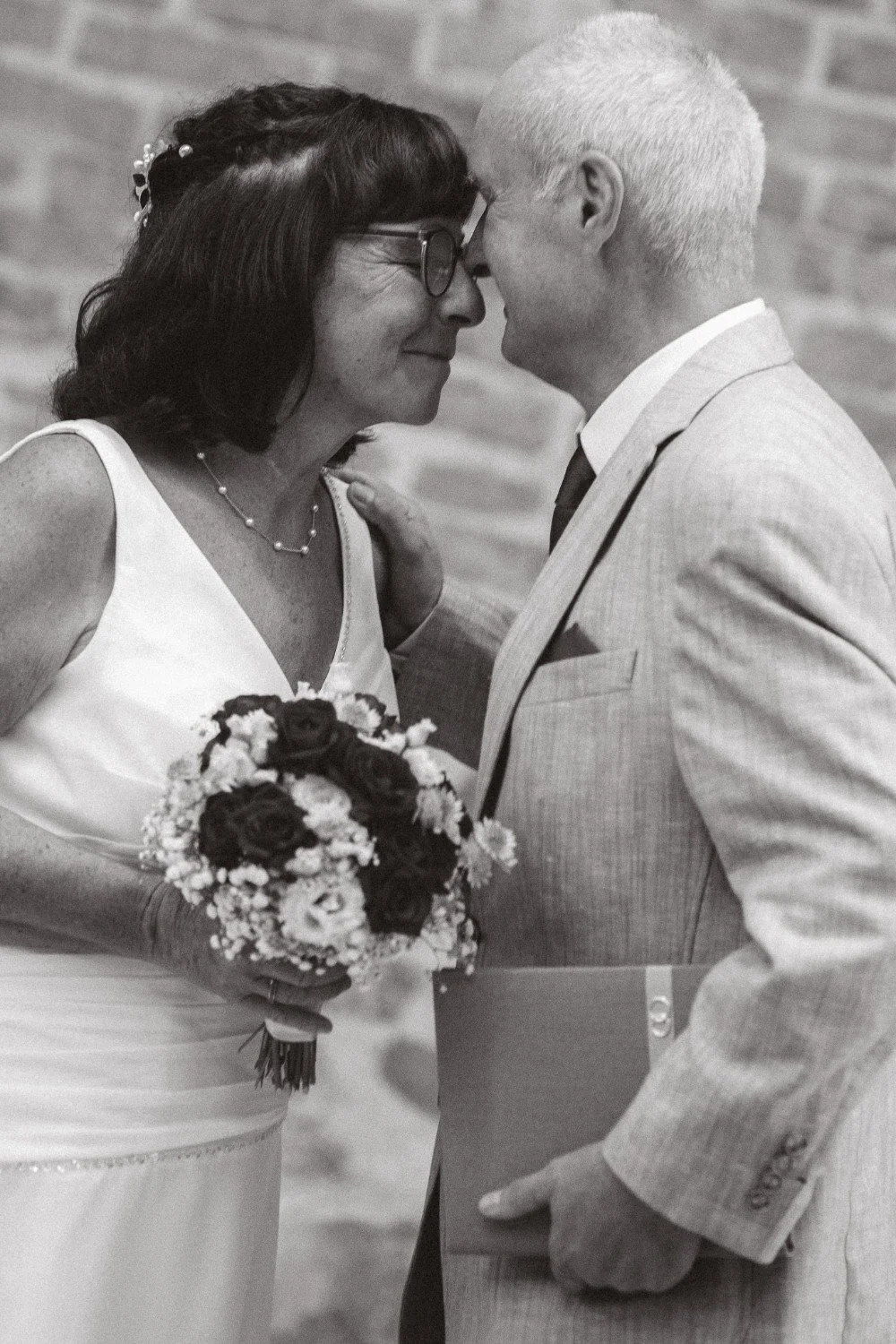 A close-up black and white portrait of the wedding couple smiling with their foreheads touching, holding a bouquet and a folder.
